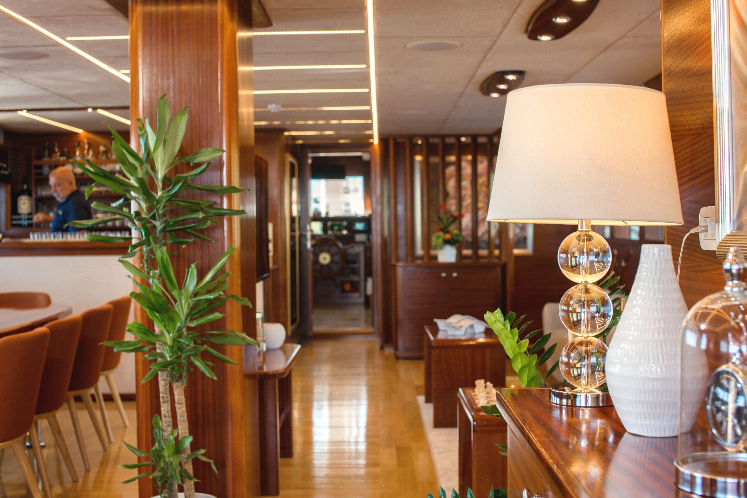 SON DE MAR A cozy, modern interior with wood-paneled walls, a potted plant, a lamp with a round glass base, and a white vase on a side table. Brown chairs are lined up near a bar, where a man stands in the background.