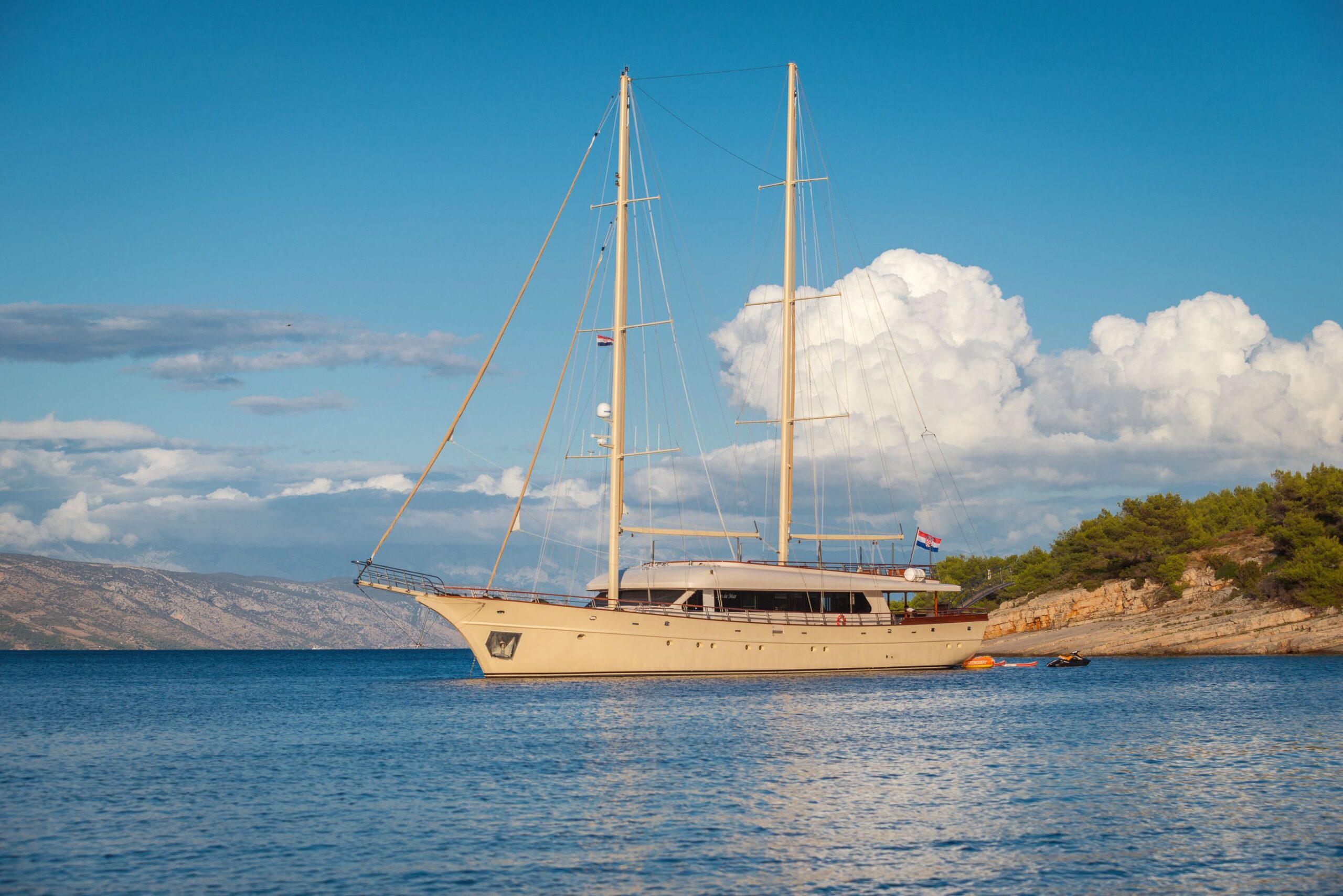 SON DE MAR A beige sailing yacht with two tall masts floats on calm blue water near a rocky, tree-lined shore under a blue sky with large white clouds.