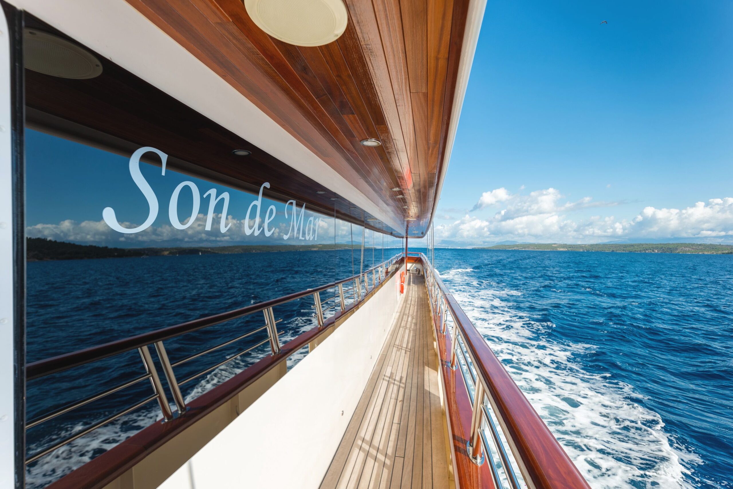 SON DE MAR View from the side deck of a boat named Son de Mar sailing on blue ocean water, with a wooden ceiling, clear sky, and distant coastline visible in the background.