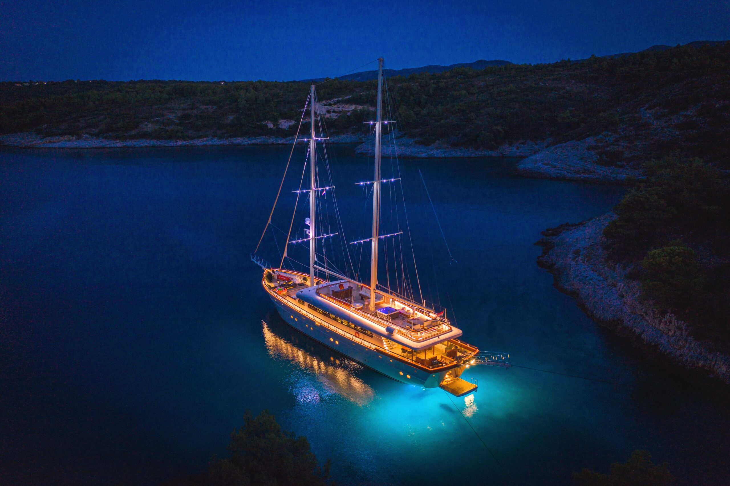 SON DE MAR A large, illuminated sailboat is anchored in calm, dark blue water near a rocky, forested shoreline at night, with underwater lights glowing beneath the vessel.