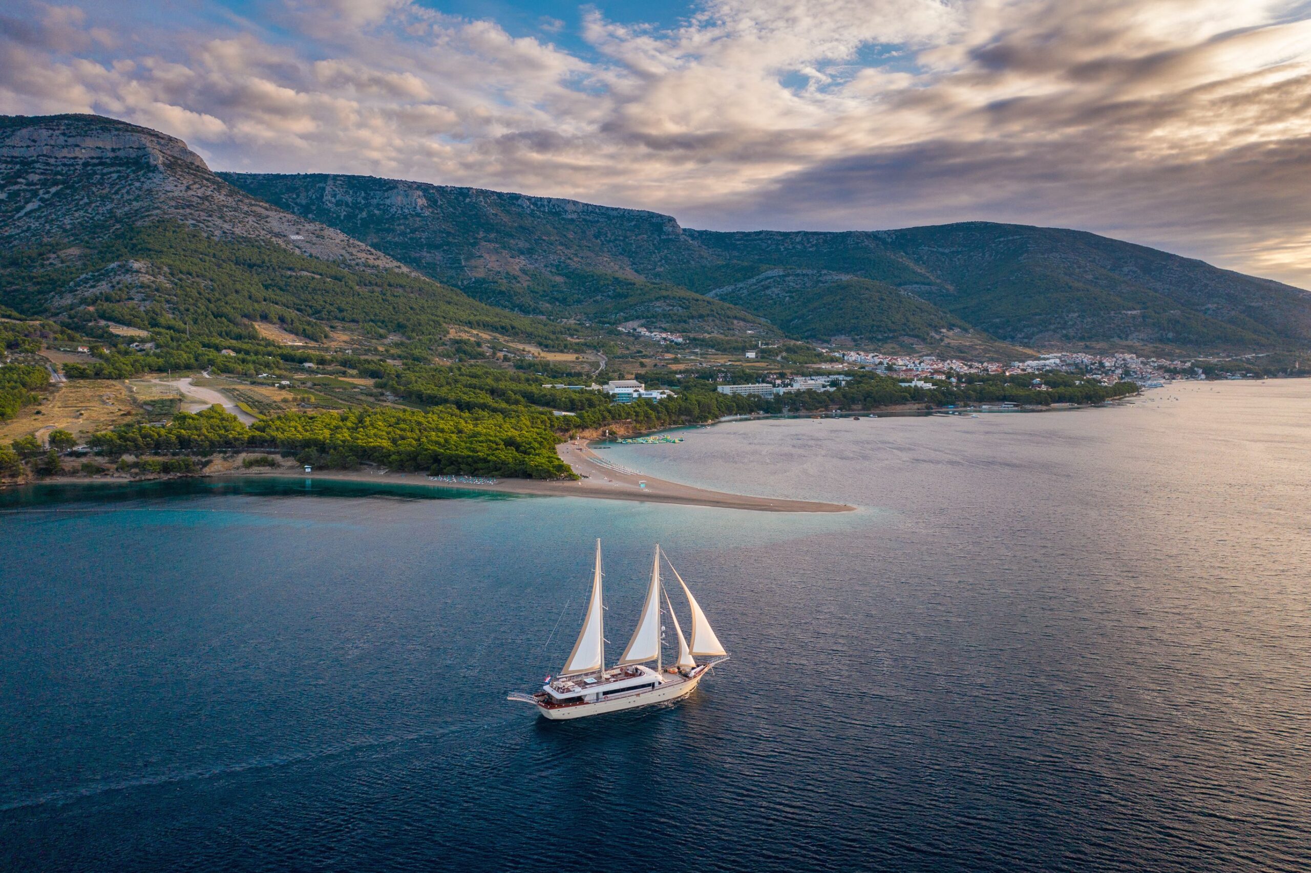 SON DE MAR A sailboat with white sails glides over deep blue water near a narrow strip of sandy beach and lush green coastline, set against mountains and a partly cloudy sky at sunset.