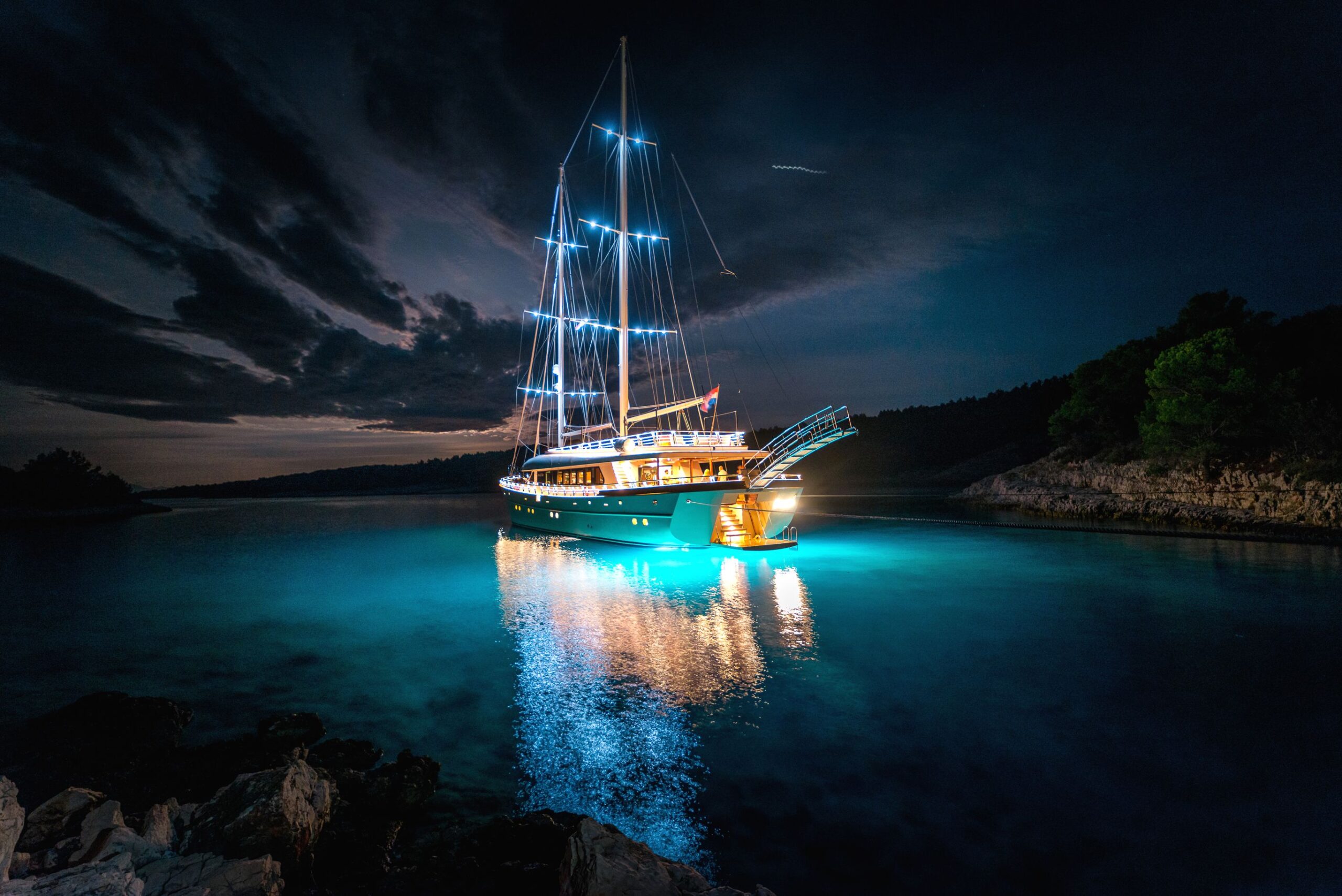 SON DE MAR A large sailboat illuminated with bright lights is anchored at night in calm water, casting glowing reflections on the blue surface. Dark clouds and trees are visible in the background.