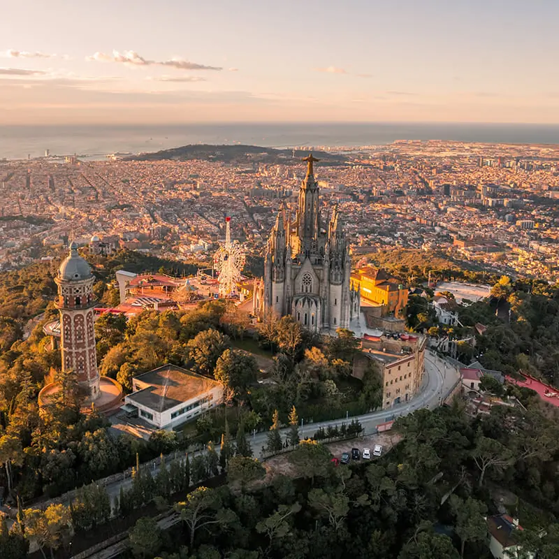 Aerial view of the Temple of the Sacred Heart of Jesus atop Tibidabo mountain, overlooking Barcelona at sunset, with city buildings and coastline in the background.