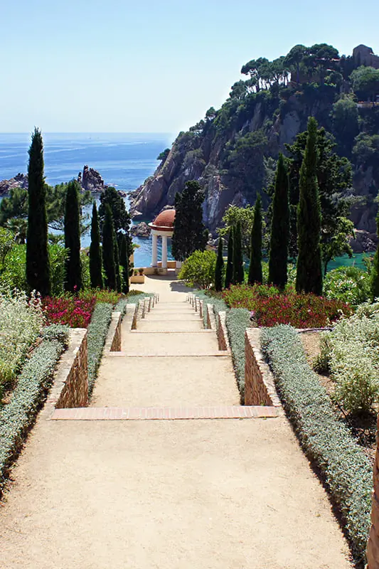 A garden path lined with tall trees and shrubs leads to a white gazebo overlooking the sea, with cliffs and blue water in the background under a clear sky.
