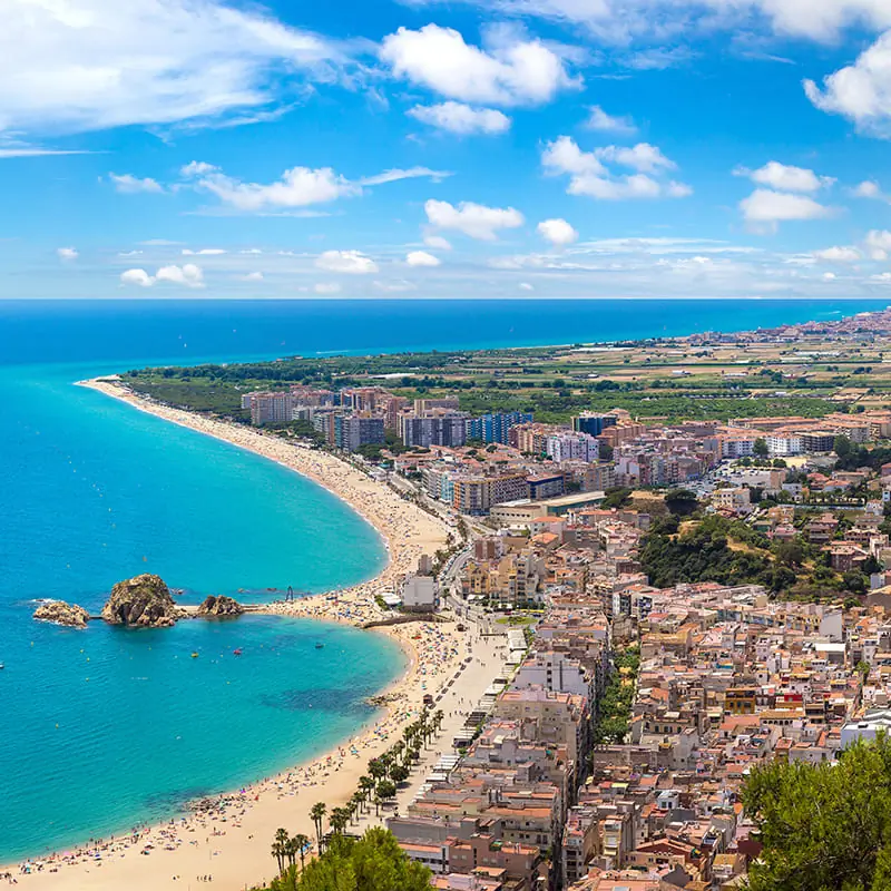 Aerial view of a coastal city with sandy beaches, blue sea, buildings, and green fields under a bright, partly cloudy sky.