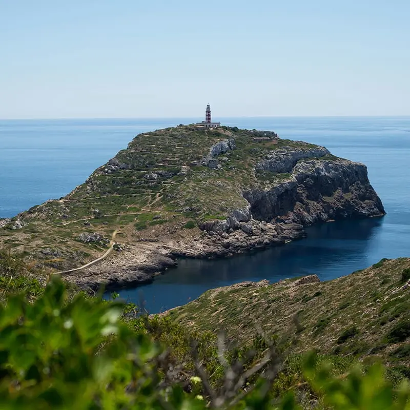 A rocky island with green vegetation juts into the blue sea, featuring a lighthouse at the highest point under a clear sky.