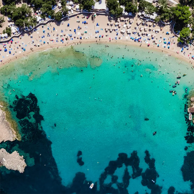 Aerial view of a crowded beach with turquoise water, swimmers, umbrellas, and rocky areas near the shore, surrounded by lush greenery.