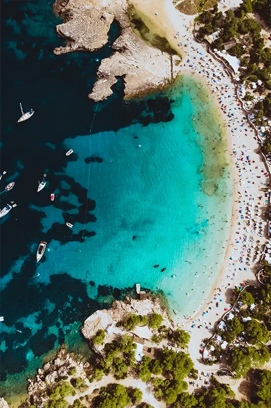 Aerial view of a turquoise bay with several boats, rocky coastline, sandy beach, and sunbathers under umbrellas surrounded by lush green trees.