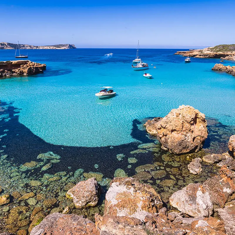 Clear turquoise water with rocky shores, several small boats floating, and a sunny blue sky. The water reveals rocks beneath the surface and a distant coastline. The scene is tranquil and picturesque, suggesting a coastal getaway.