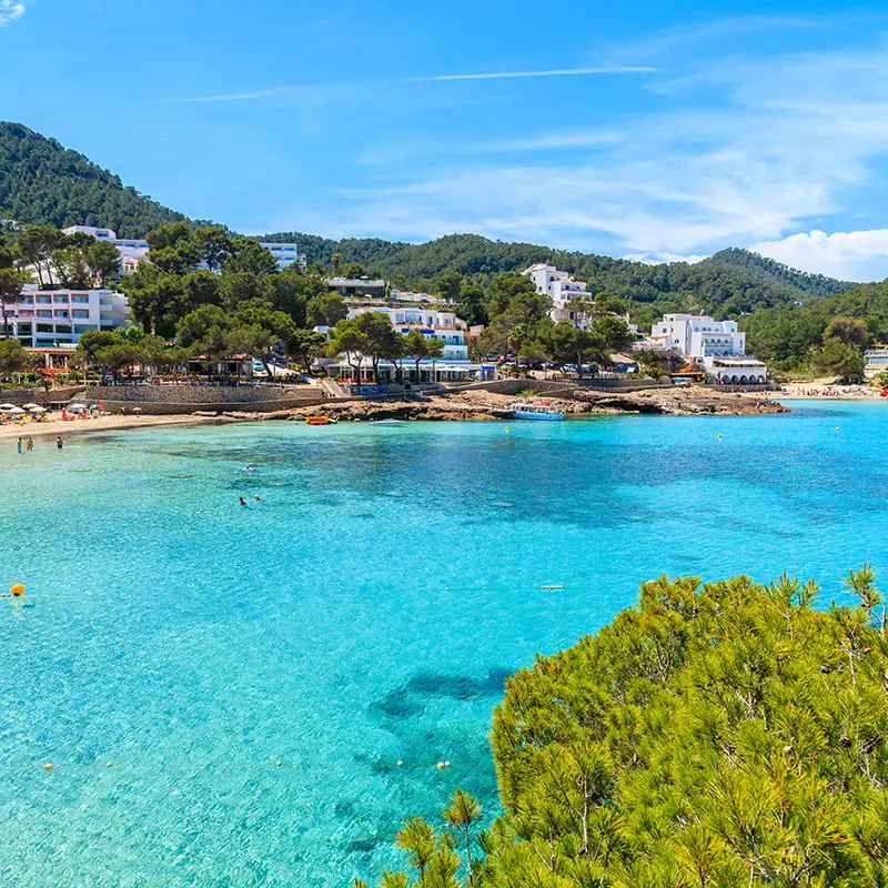 A clear turquoise bay with a sandy beach, surrounded by green trees and white buildings on a hillside under a blue sky with wispy clouds.