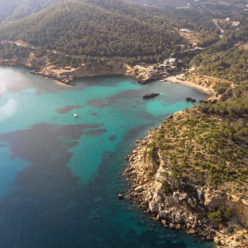 Aerial view of a rocky coastline with clear turquoise water, a small boat in the bay, and forested hills in the background.