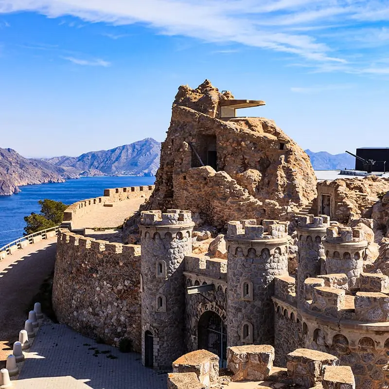 A stone castle-like structure with round towers sits by a winding road, overlooking a blue sea and distant rugged mountains under a bright sky.