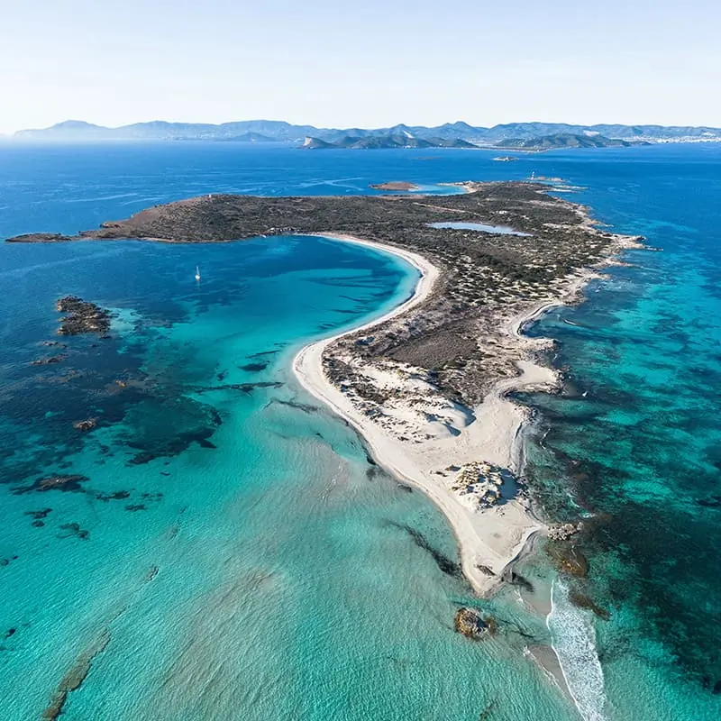 Aerial view of a narrow island with white sandy beaches, turquoise waters, and rocky areas, surrounded by a calm, blue sea under a clear sky.