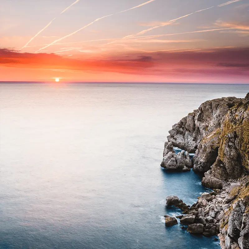Rocky cliff by a calm sea at sunset, with the sun partially visible on the horizon and streaks of clouds in the colorful sky.
