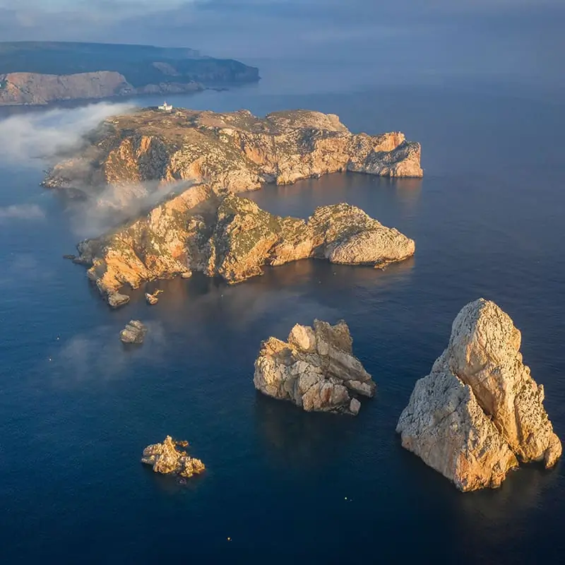 Aerial view of rocky islands surrounded by blue sea, with mist drifting over the land and cliffs in the background under a partly cloudy sky.