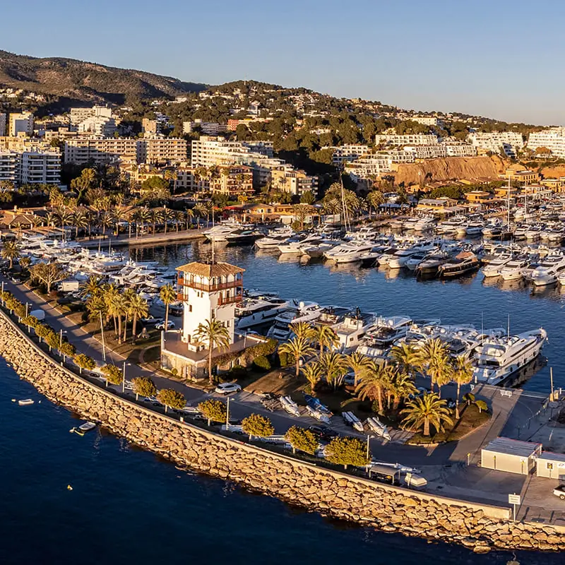 Aerial view of a marina filled with yachts, a waterfront promenade lined with palm trees, and a small white tower, with a coastal town and hills in the background under a clear sky.