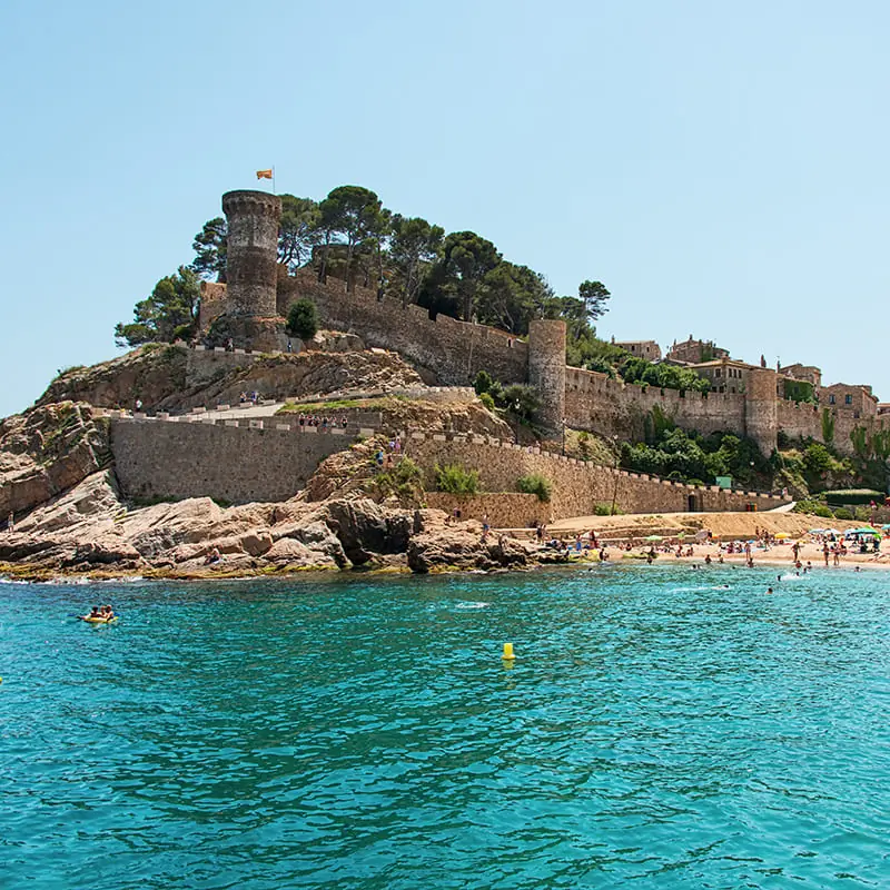 A stone castle with towers sits atop a rocky hill, overlooking a sandy beach and turquoise sea, with people swimming and relaxing in the sun.
