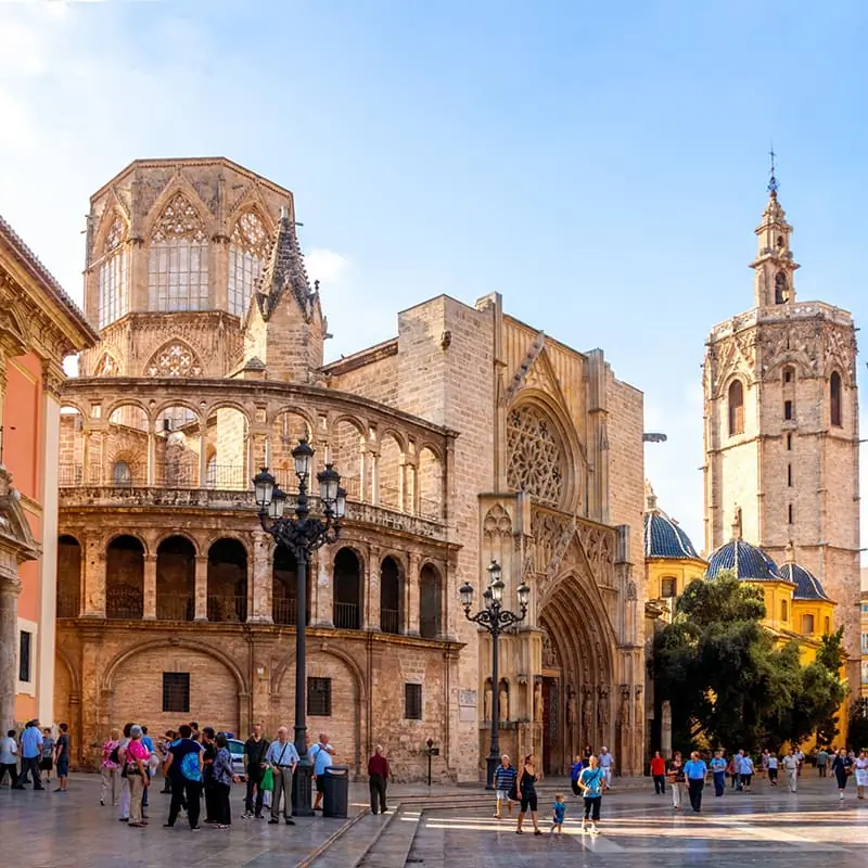 A group of people walk and gather near a large historic cathedral with ornate stone architecture and a tall bell tower under a sunny blue sky.