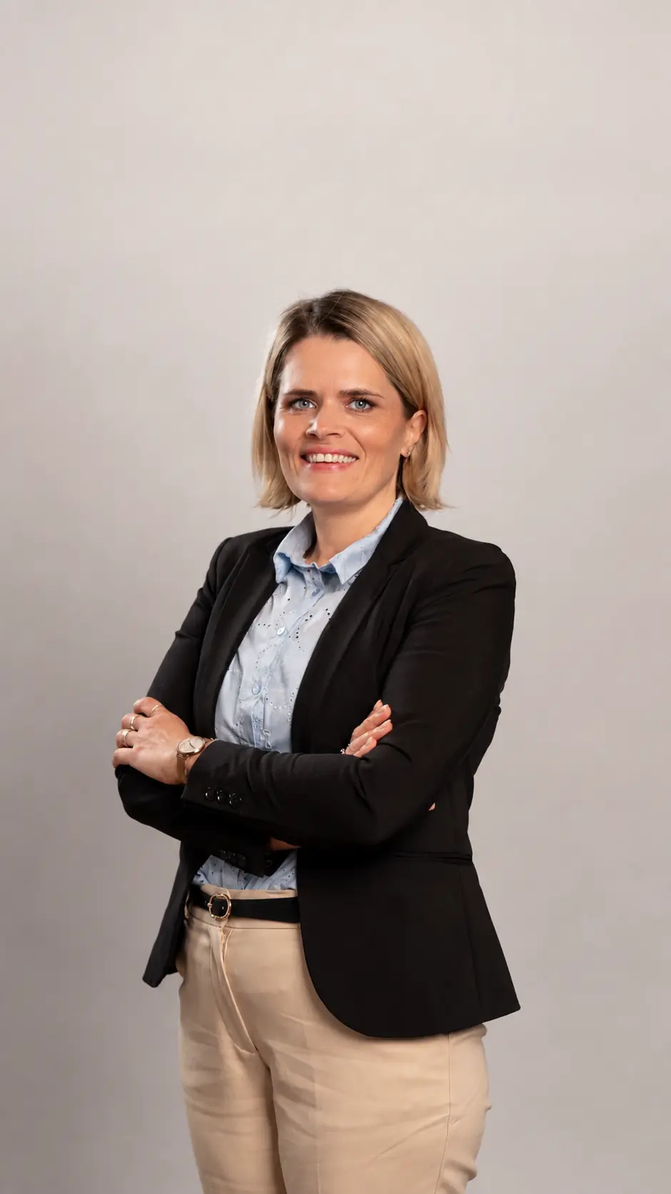 Woman with short blonde hair wearing a black blazer, light blue shirt, and beige pants, stands smiling with arms crossed in front of a plain light gray background.