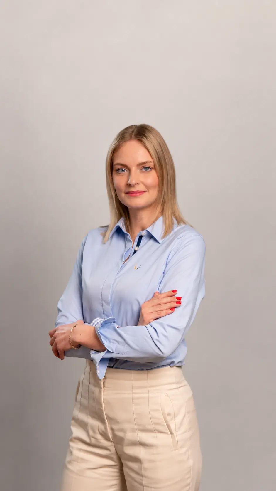 A woman with blonde hair wearing a light blue button-up shirt and beige pants stands confidently with arms crossed, smiling, against a plain light gray background.