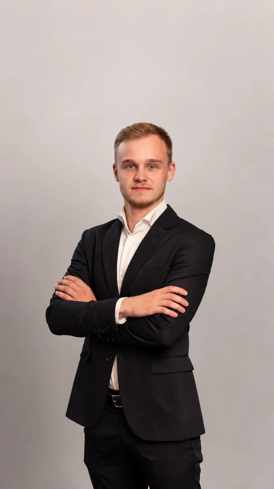 Young man in a black suit with a white shirt stands against a plain light gray background, facing forward with arms crossed and a neutral expression.