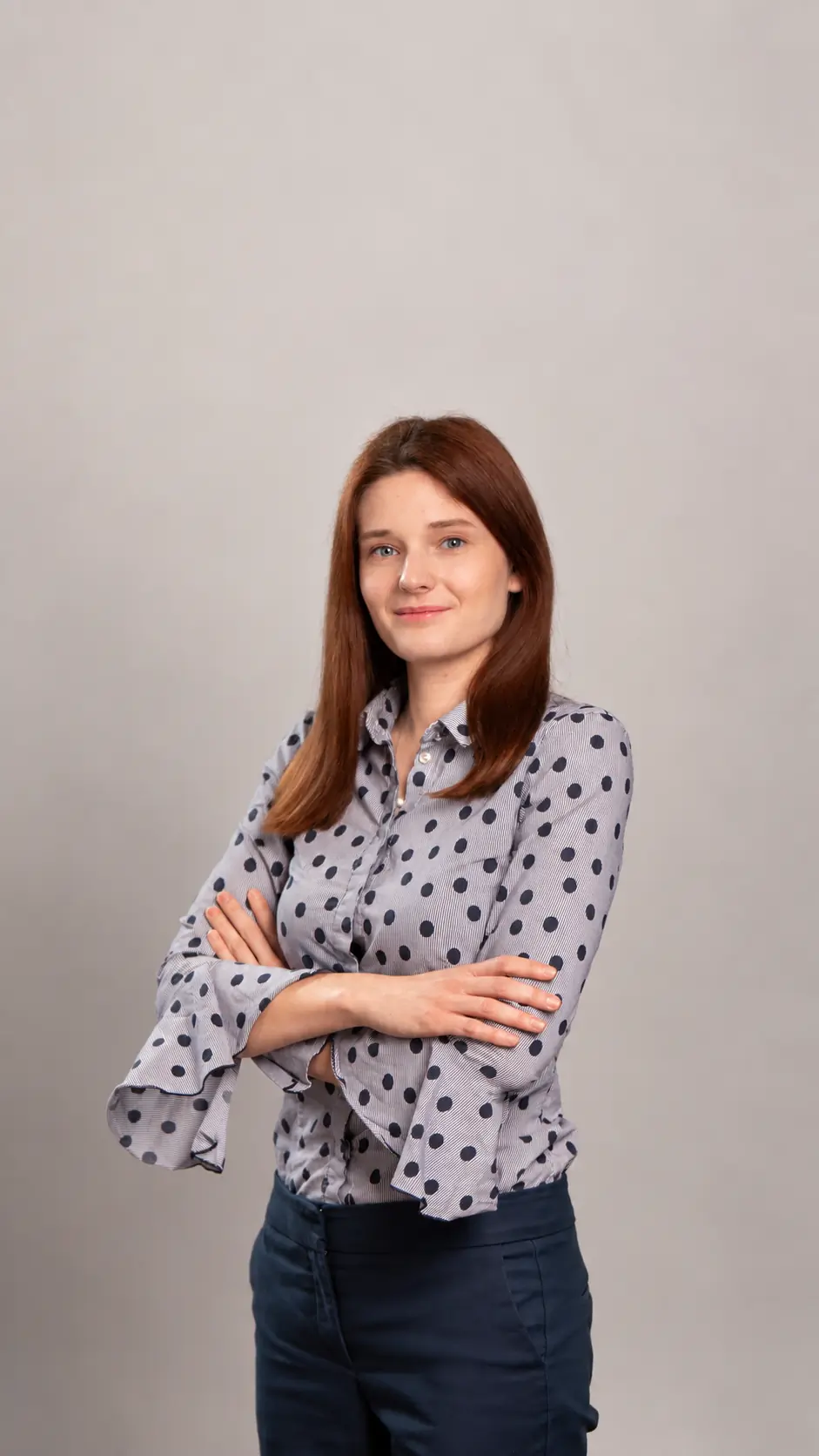A woman with straight brown hair stands against a plain background, wearing a gray blouse with black polka dots and navy pants. She is smiling slightly and has her arms crossed.