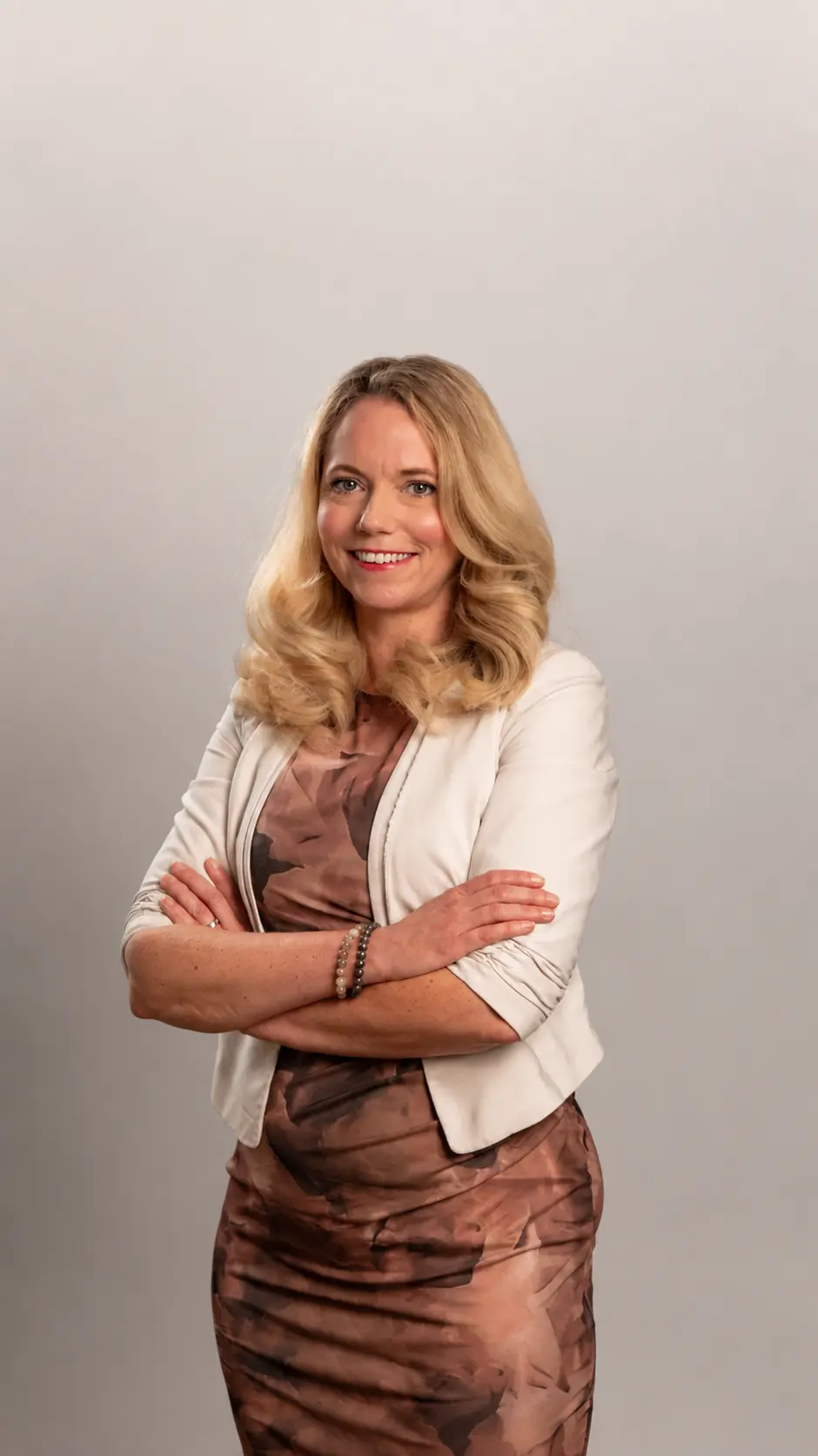 A woman with long blonde hair, wearing a light blazer over a patterned dress, stands with arms crossed and smiles against a plain light background.
