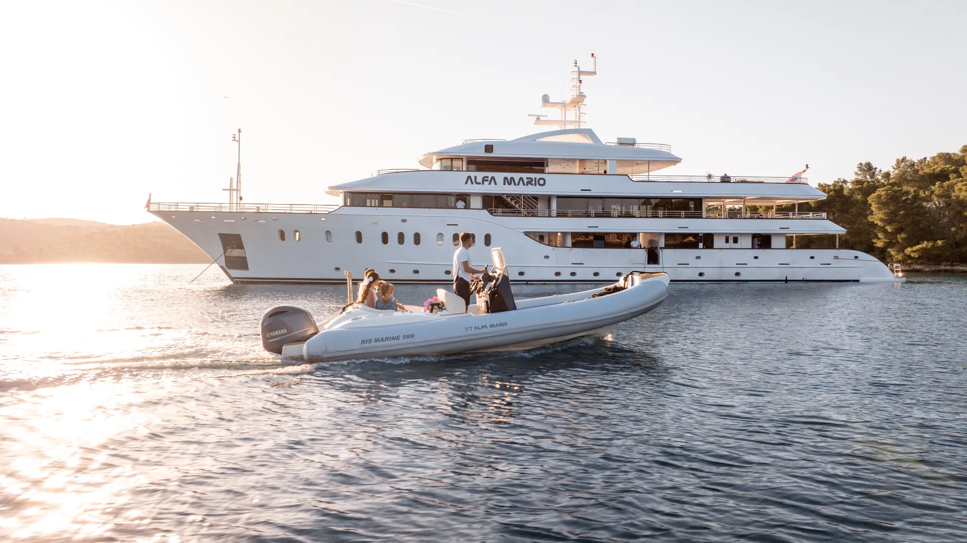 A large white yacht named Alfa Mario is anchored on calm water, with several people riding a smaller inflatable boat in the foreground during a sunny day. Trees and hills are visible in the background.