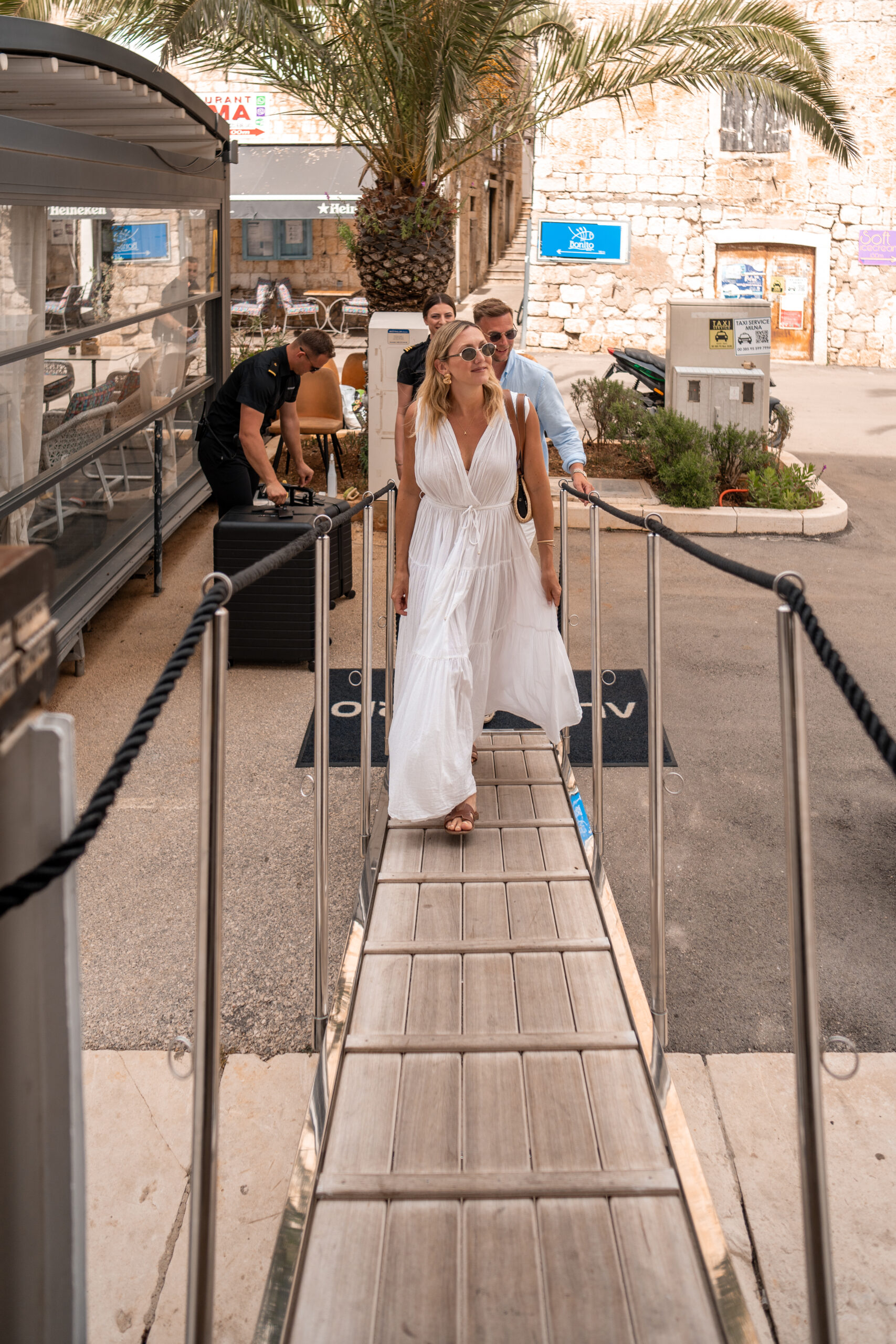 A woman in a flowing white dress and sunglasses walks down a dock ramp, followed by a man and another woman, with palm trees and stone buildings in the background.
