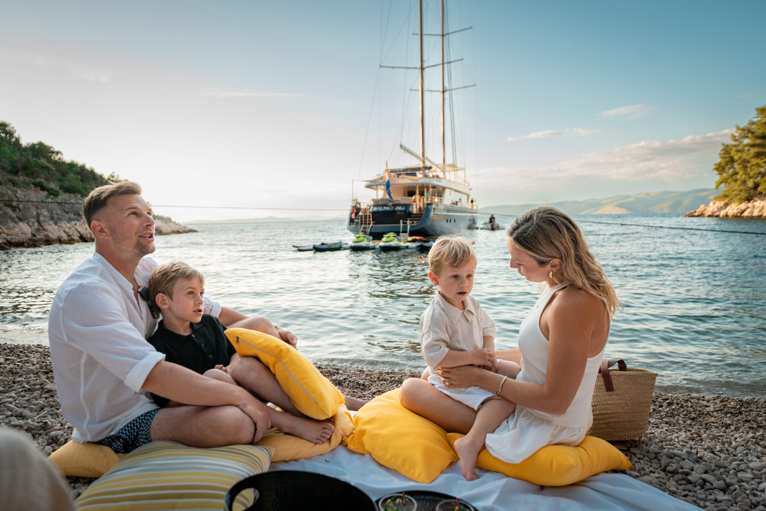 A family of four relaxes on a rocky beach with yellow cushions and a picnic, with a sailboat anchored in the calm water behind them on a sunny day.