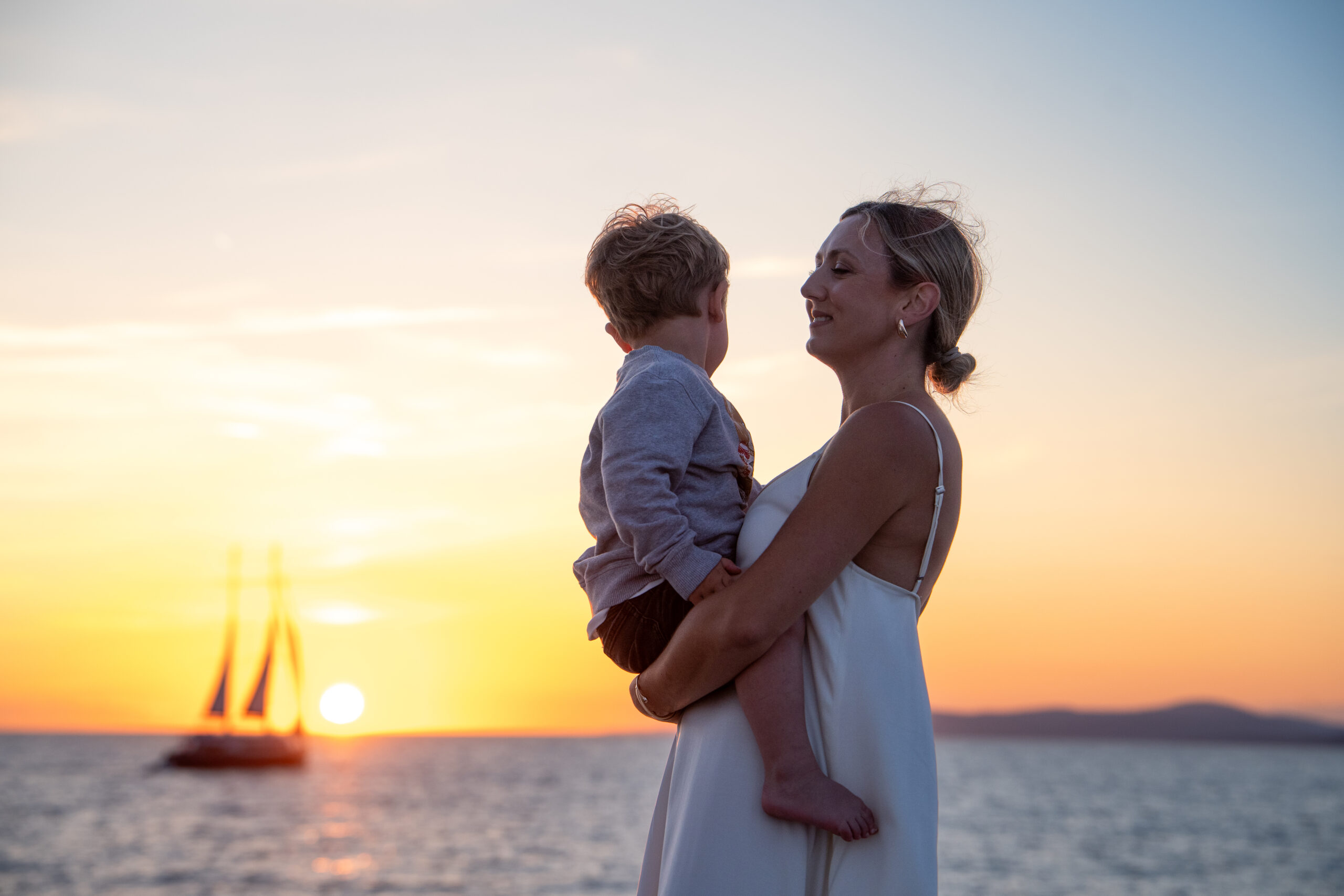 A woman in a white dress holds a young child by the ocean at sunset; a sailboat is visible on the water with the sun low on the horizon.