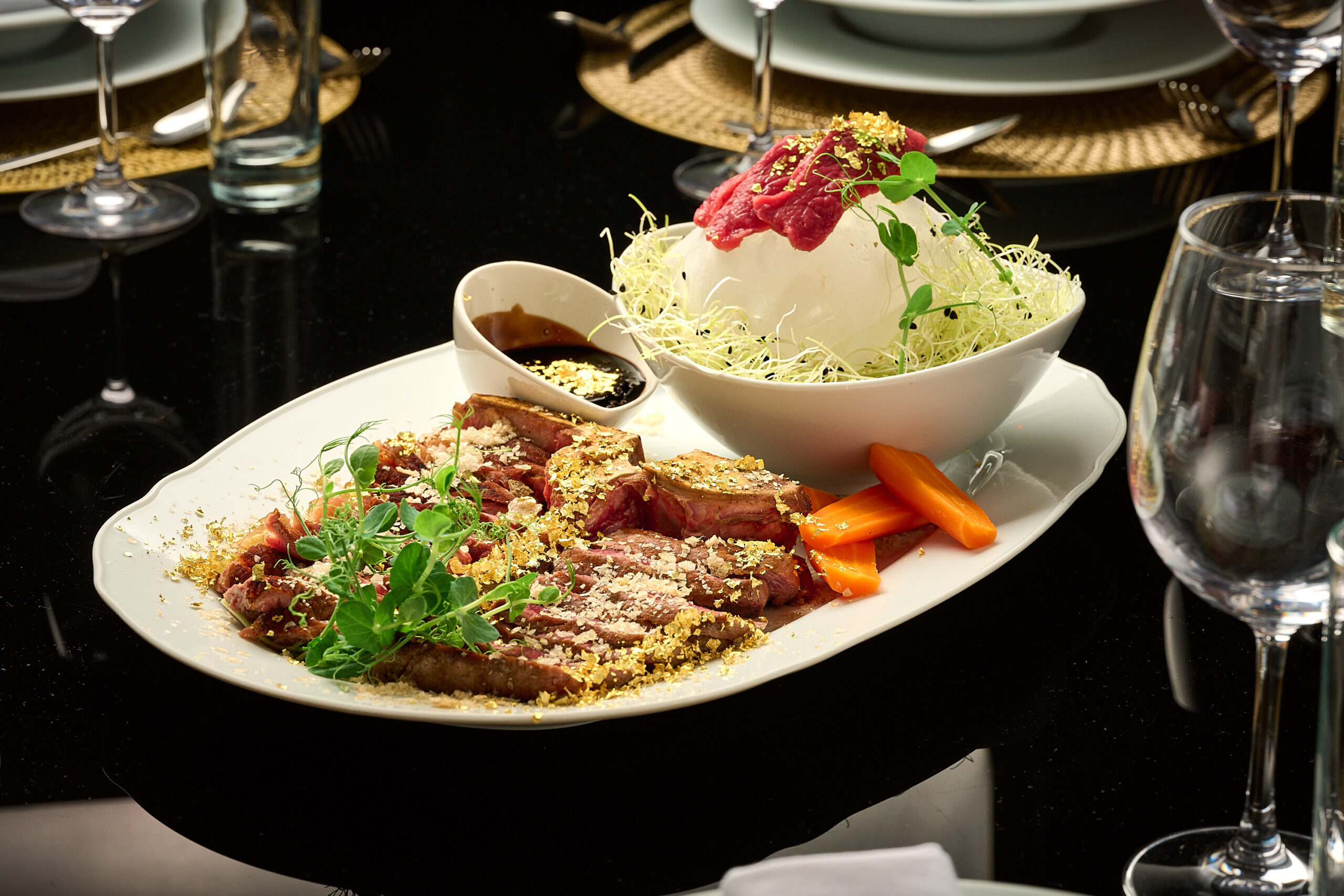 A white plate with sliced steak topped with herbs and crumbs, served with carrot sticks, sauce, and a bowl of shredded cabbage salad garnished with sprouts on a black table set with glassware and plates.