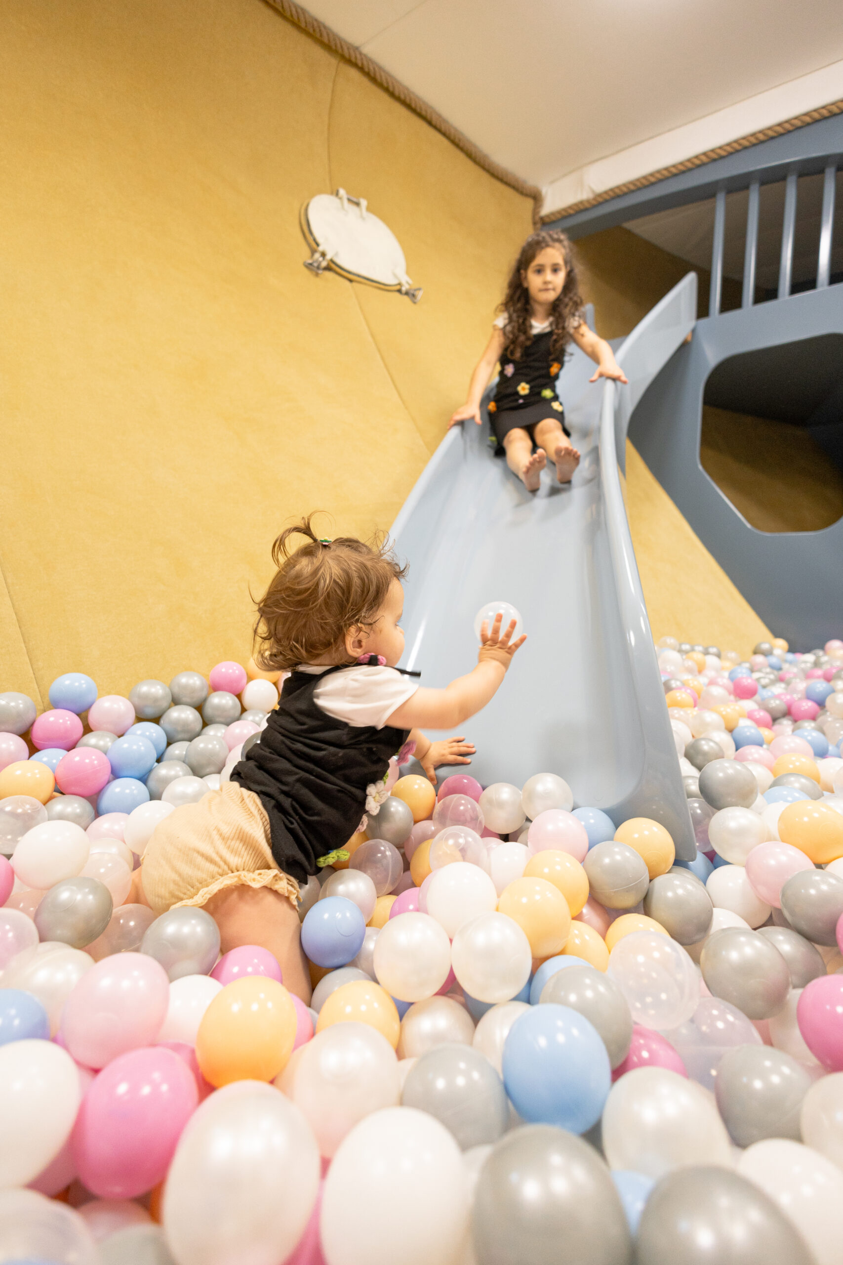 Two young children play in a ball pit; one slides down a gray slide while the other reaches for a white ball among pastel-colored balls on the floor. The setting appears to be an indoor play area.