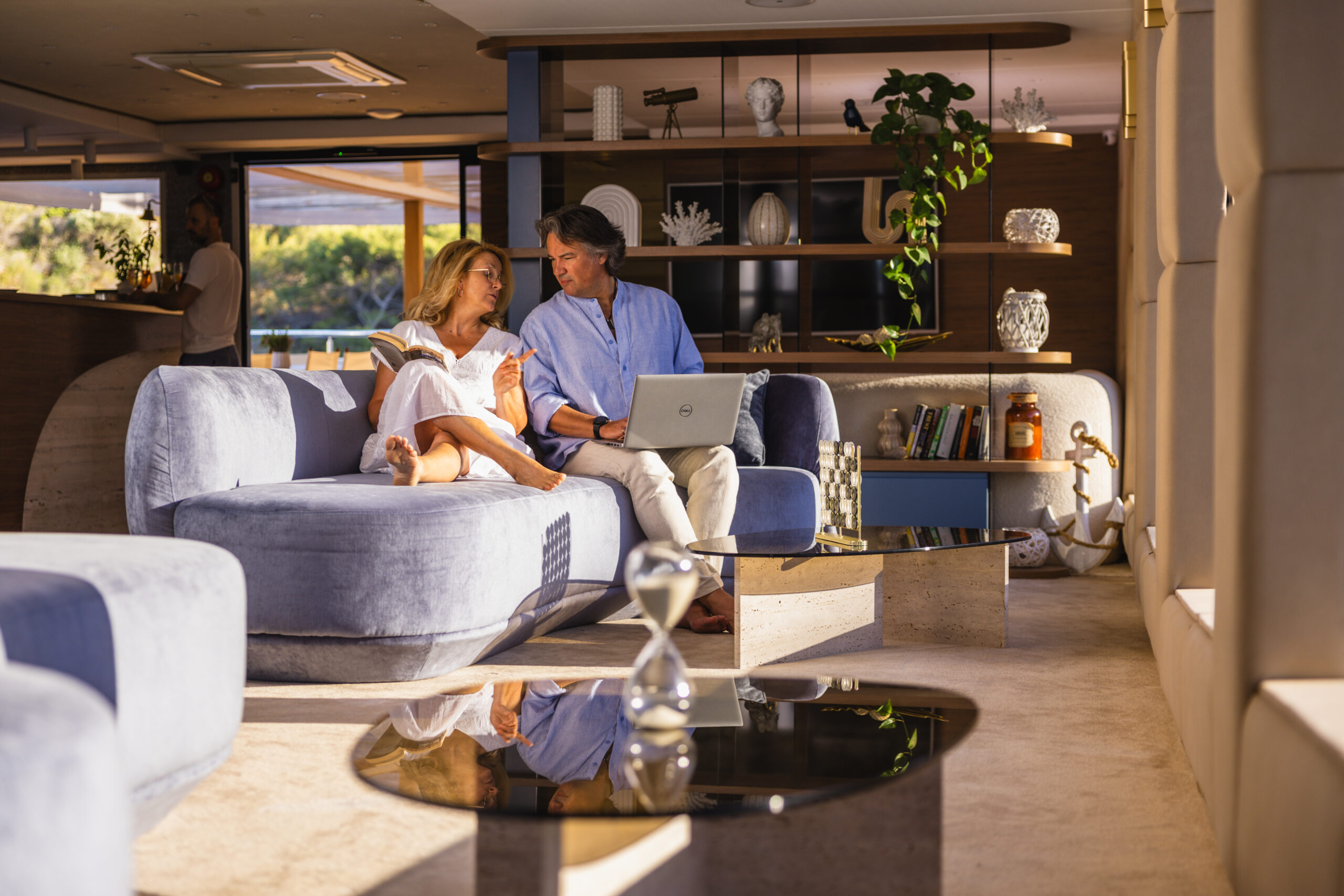 A couple relaxes on a modern, stylish sofa indoors. The woman reads a book while the man uses a laptop. Shelves with books, plants, and decor fill the background, and sunlight streams in through large windows.
