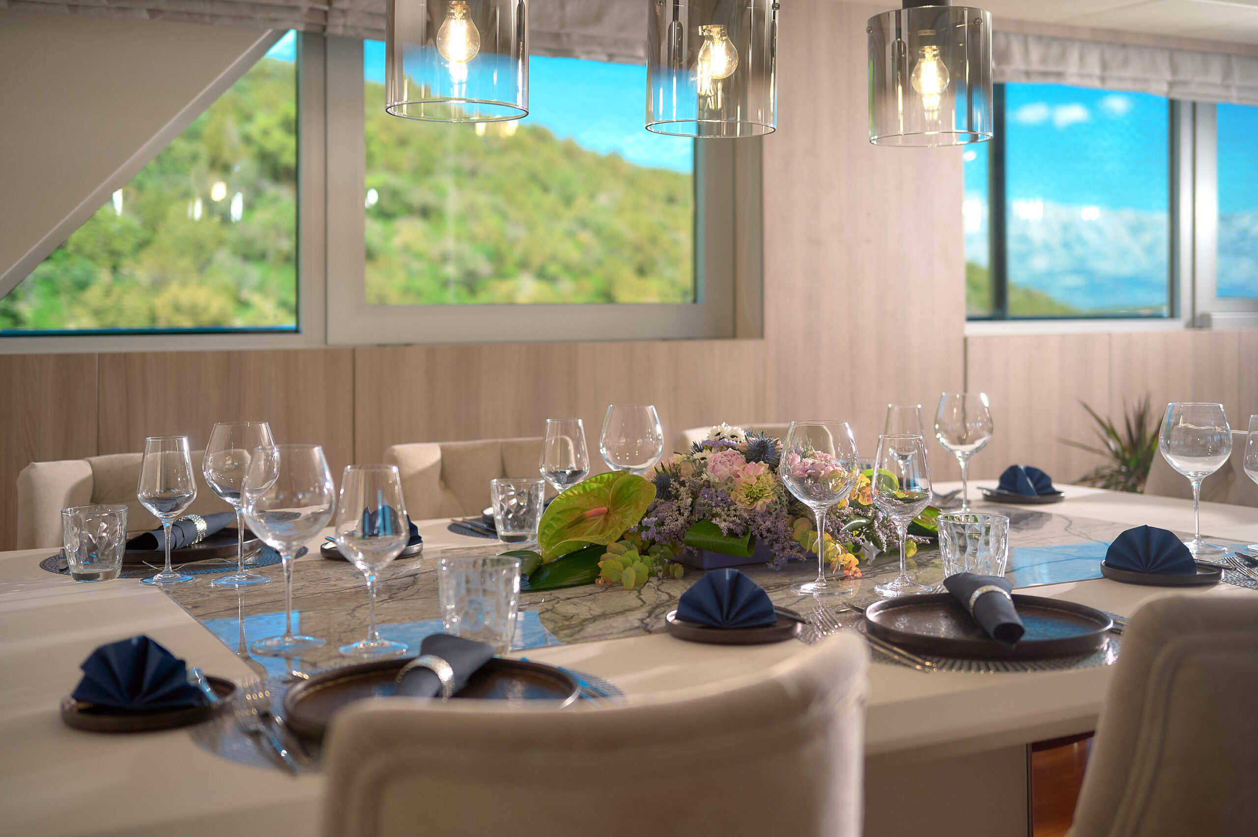 Elegant dining table set for a meal with folded navy napkins, glasses, and plates. A floral centerpiece decorates the middle. Large windows offer a view of green hills and blue sky in the background.