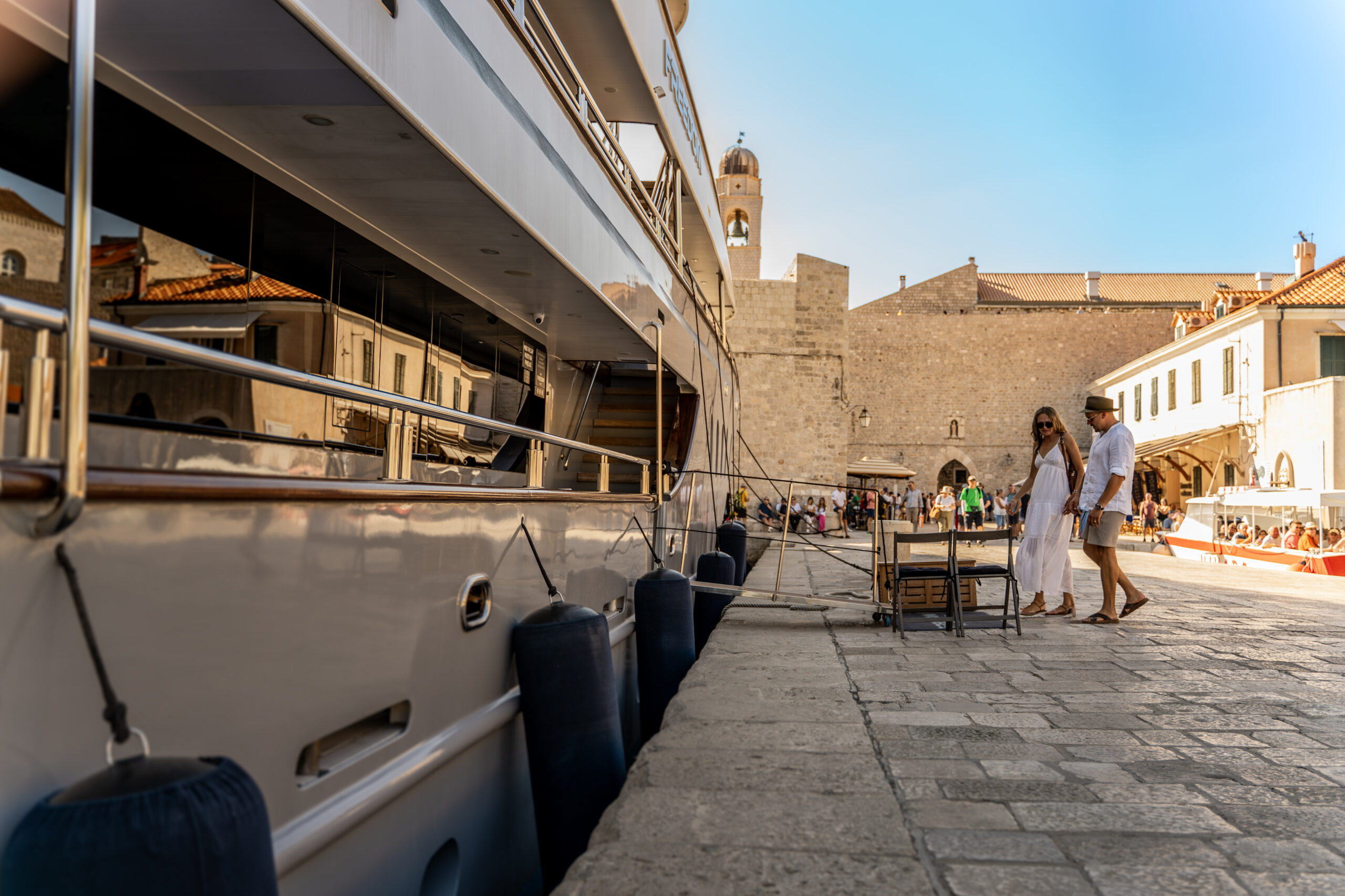 A couple walks along a stone dock beside a large yacht on a sunny day, with historic buildings and a clock tower visible in the background.
