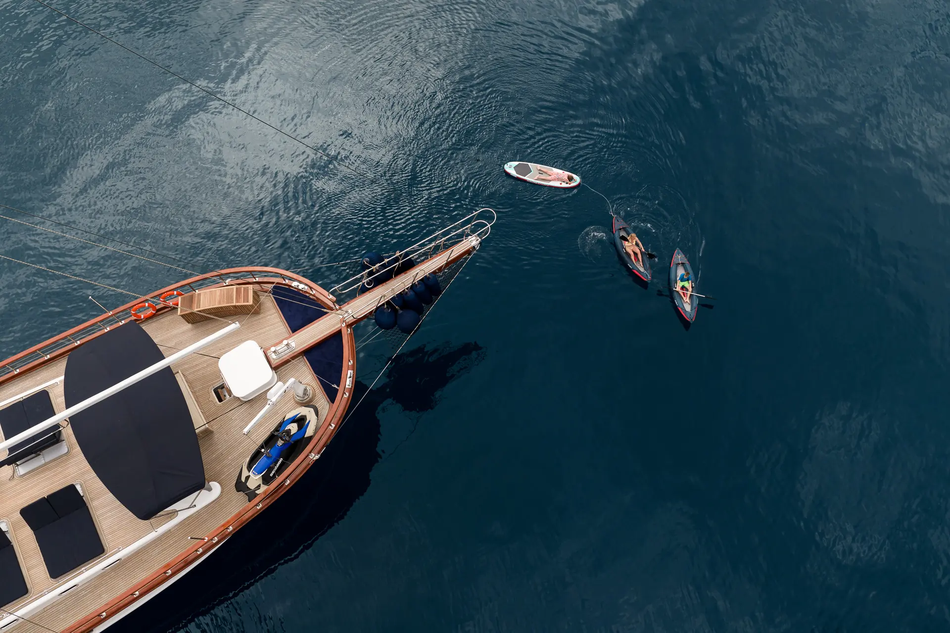Aerial view of a yacht anchored on calm, deep blue water, with two people kayaking and one person paddleboarding nearby.
