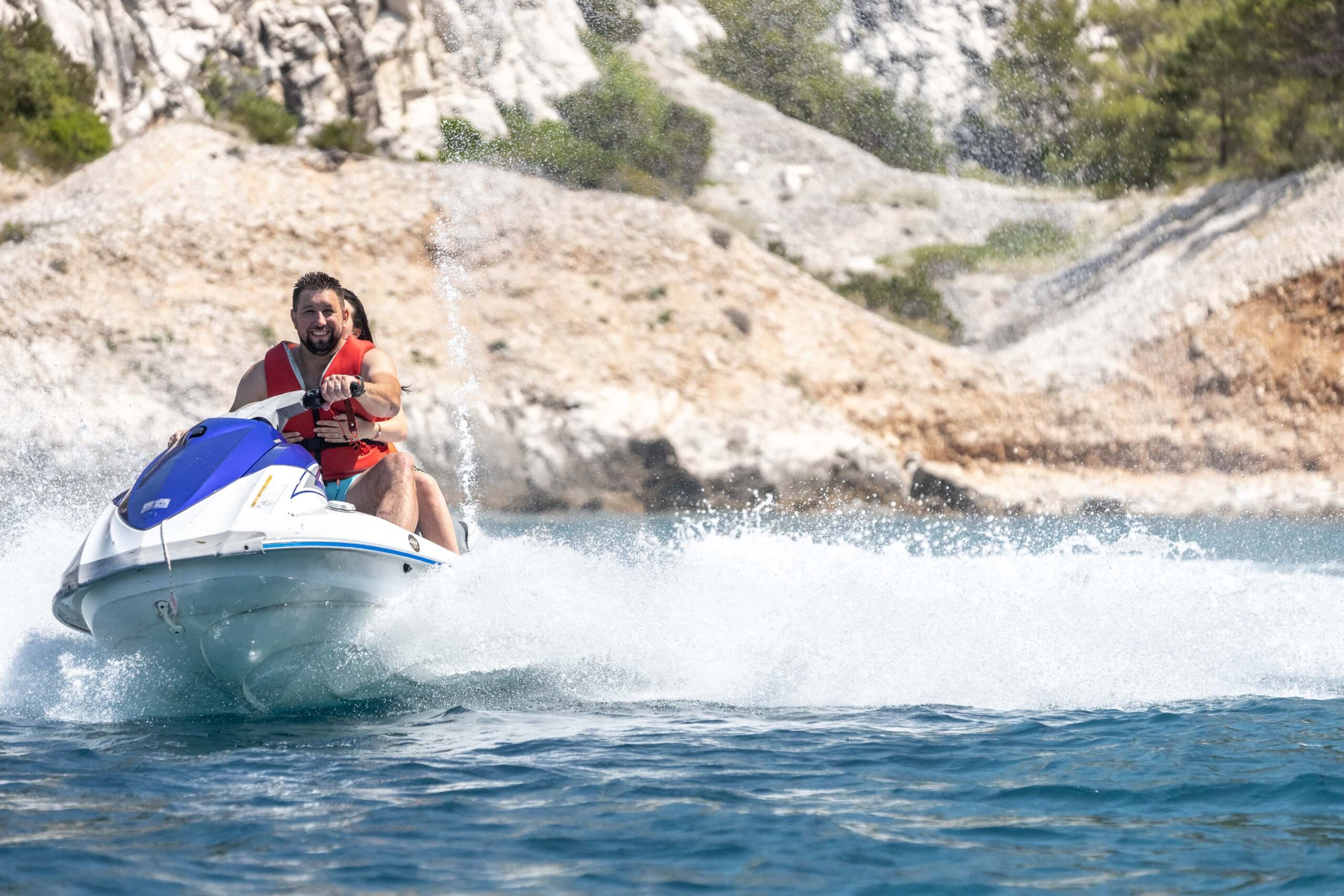 A man wearing a red life jacket rides a blue and white jet ski at high speed, creating a large splash, with rocky cliffs and greenery in the background.
