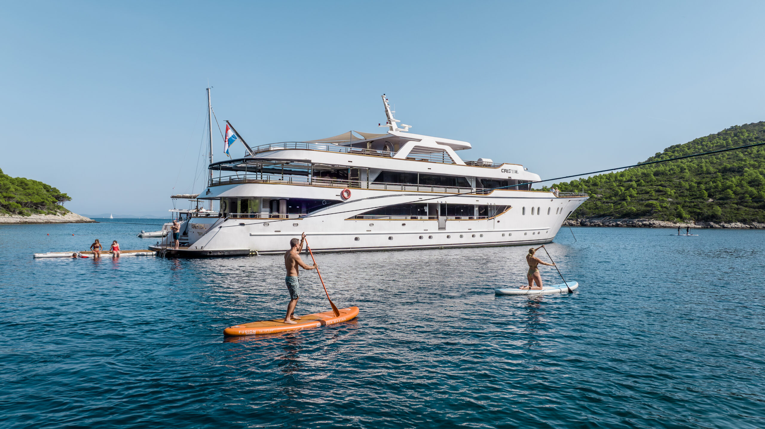 A large white yacht is anchored in calm blue water near a forested shoreline. People are paddleboarding and swimming around the yacht under a clear, sunny sky.