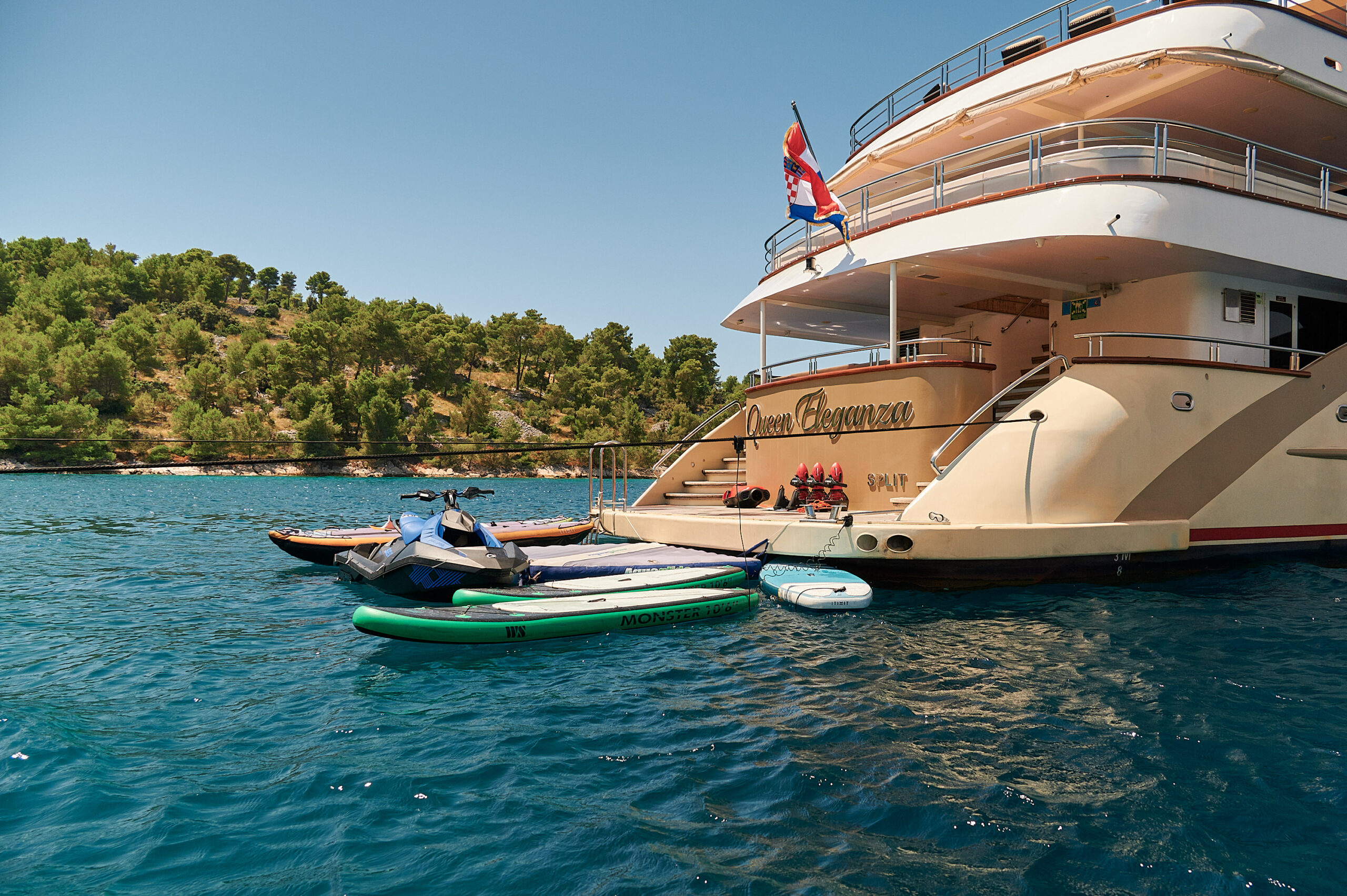A luxury yacht anchored near a wooded shoreline with jet skis, paddleboards, and a small boat floating alongside in clear blue water. A flag is flying from the yacht’s upper deck.