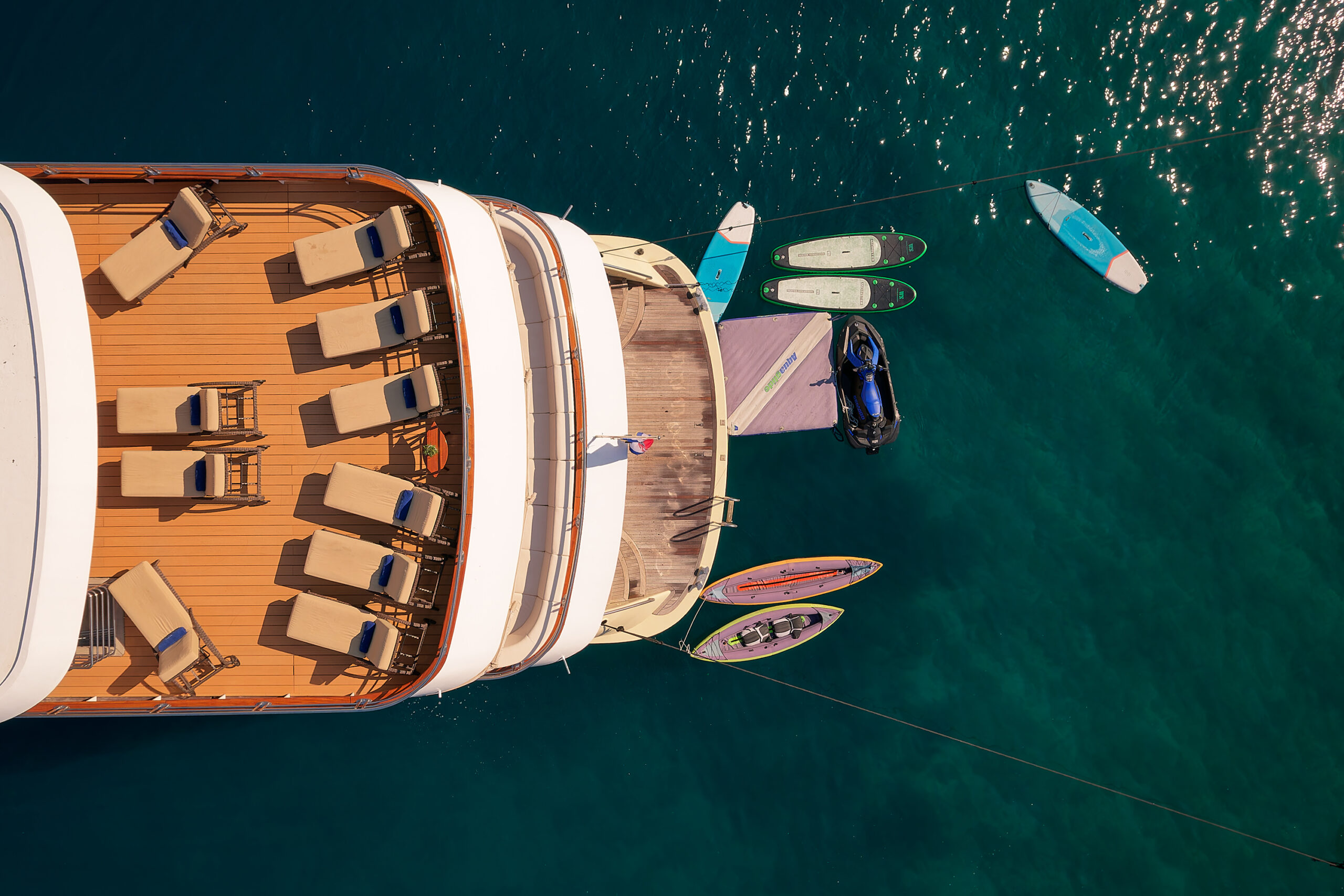 Aerial view of a yacht deck with lounge chairs, a small dock, several paddleboards, and a jet ski floating on clear blue-green water.