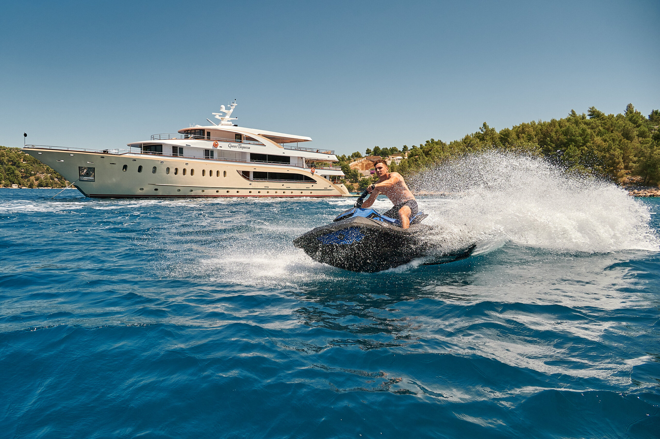 A man rides a jet ski across blue ocean water, creating a large spray, with a luxurious yacht and forested shoreline visible in the background under a clear sky.