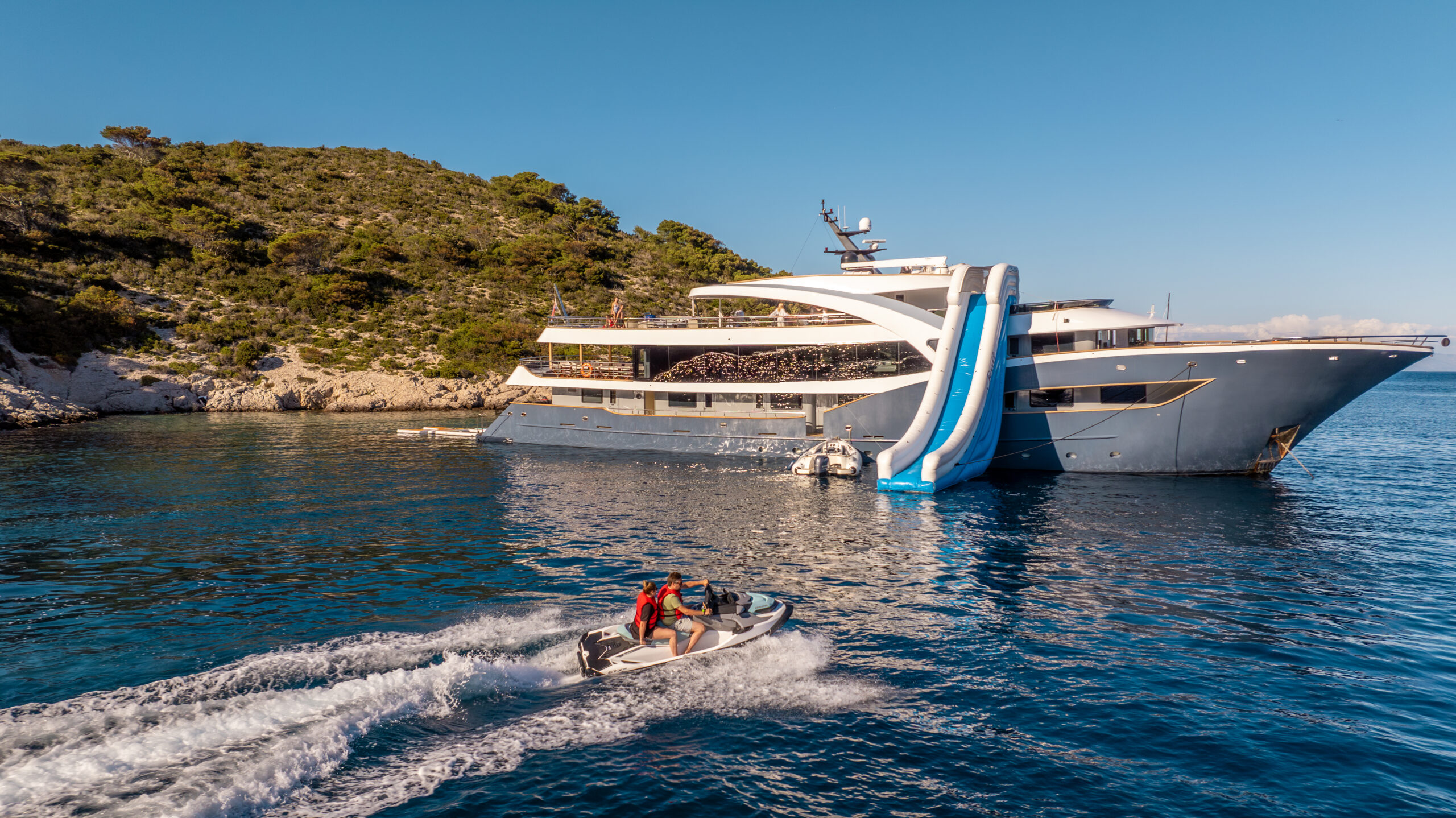 A large luxury yacht with a blue and white waterslide is anchored near a rocky, green shoreline. Two people ride a jetski on the calm blue water in the foreground, leaving a trail behind them.