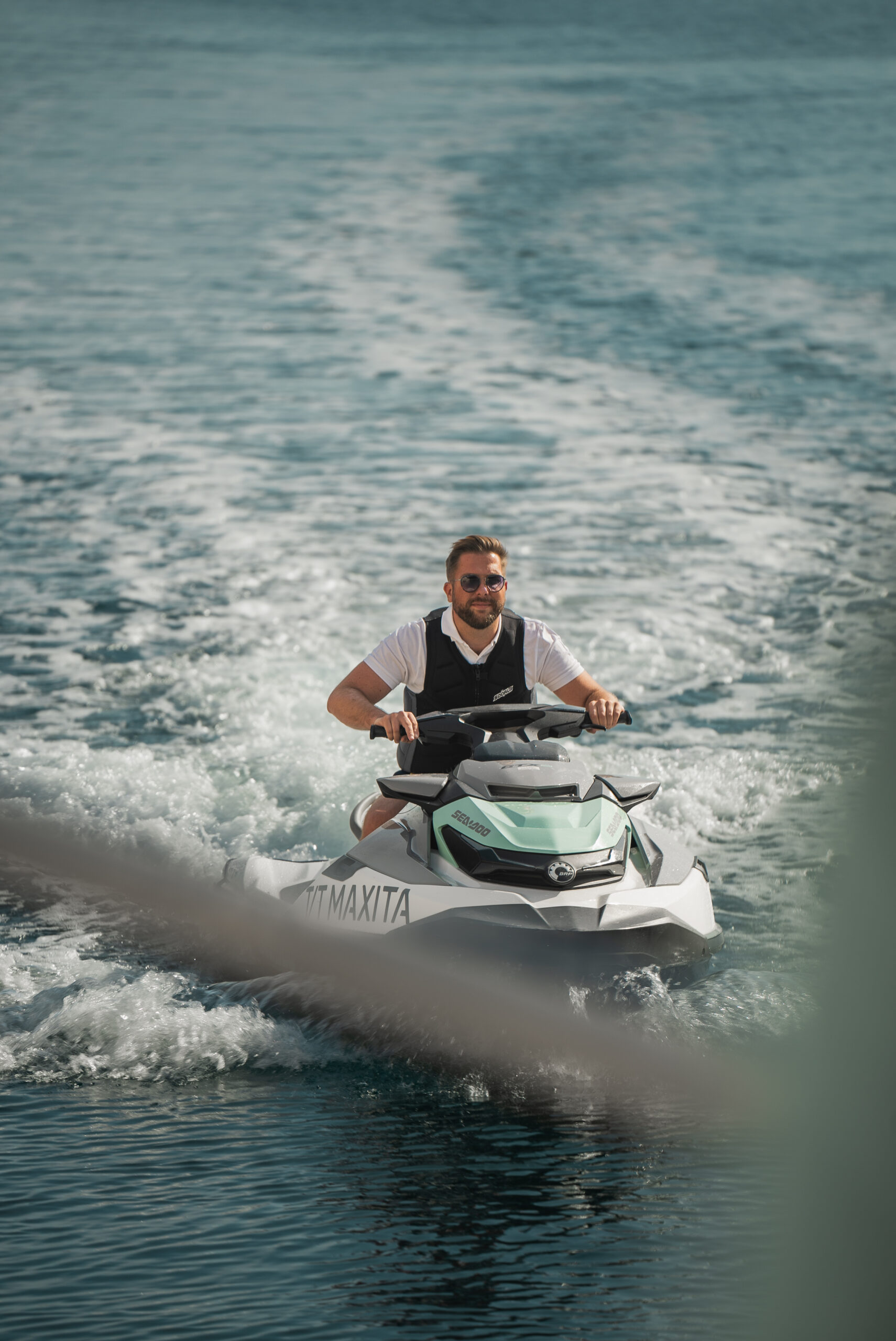 A man wearing sunglasses and a life vest rides a white jet ski on clear blue water, creating a trail of waves behind him under a sunny sky.