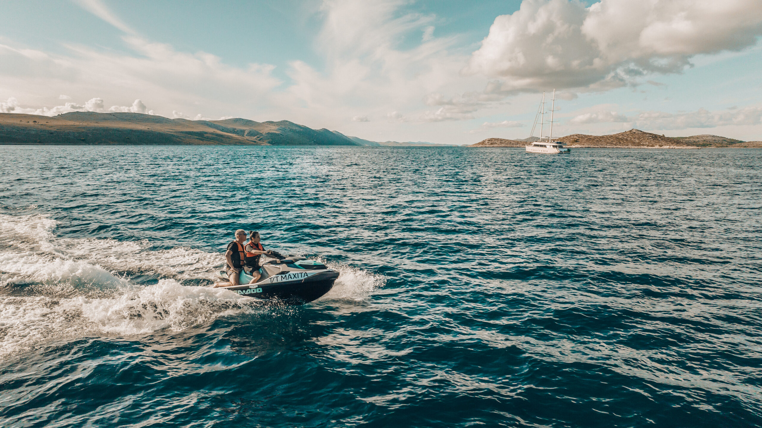 Two people ride a jet ski across deep blue water under a partly cloudy sky, with hills and a sailboat visible in the distance.