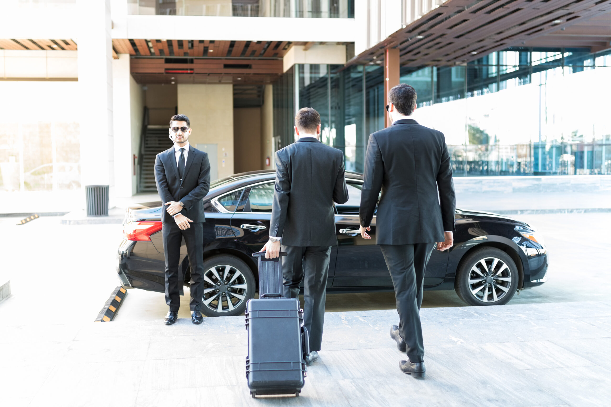 Three men in suits, one holding a suitcase, walk toward a black car parked outside a modern building; another man in a suit stands beside the car, appearing to wait or guard.