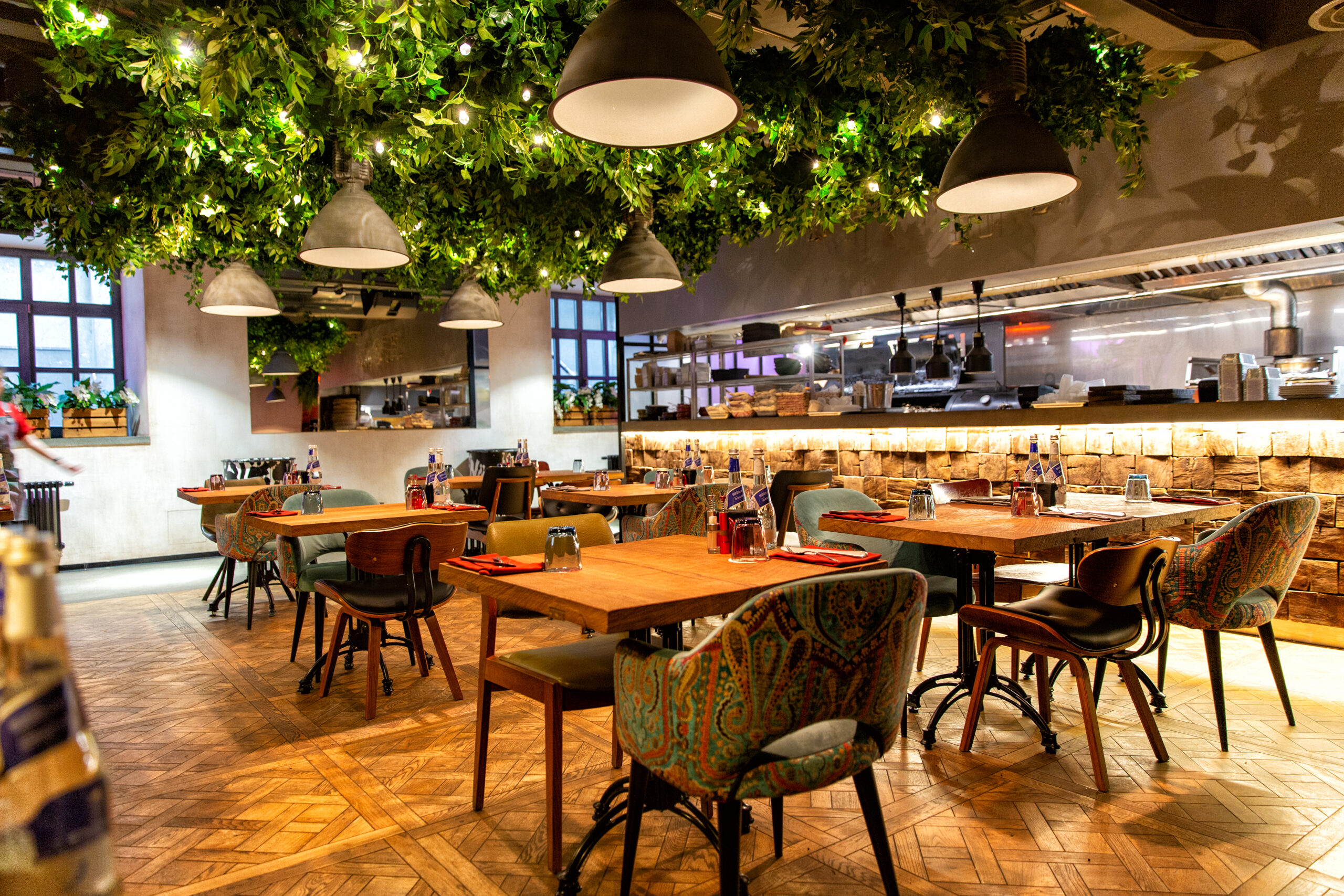 A modern restaurant interior with wooden tables, colorful patterned chairs, hanging pendant lights, and a ceiling decorated with green foliage and string lights. The open kitchen is visible in the background.
