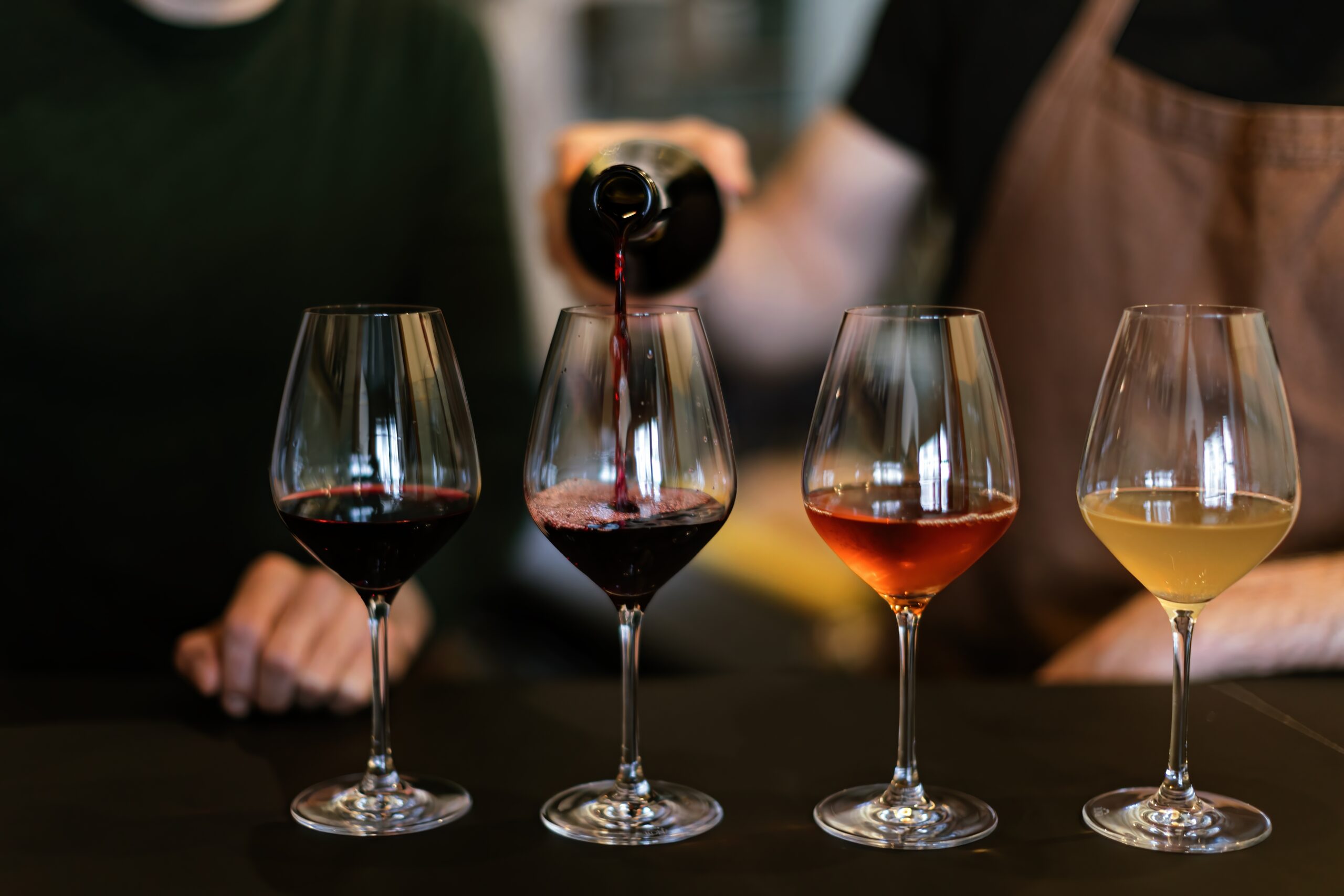 A person in an apron pours red wine into one of four wine glasses on a table, each glass containing a different type or color of wine. Another person sits nearby, partially visible.