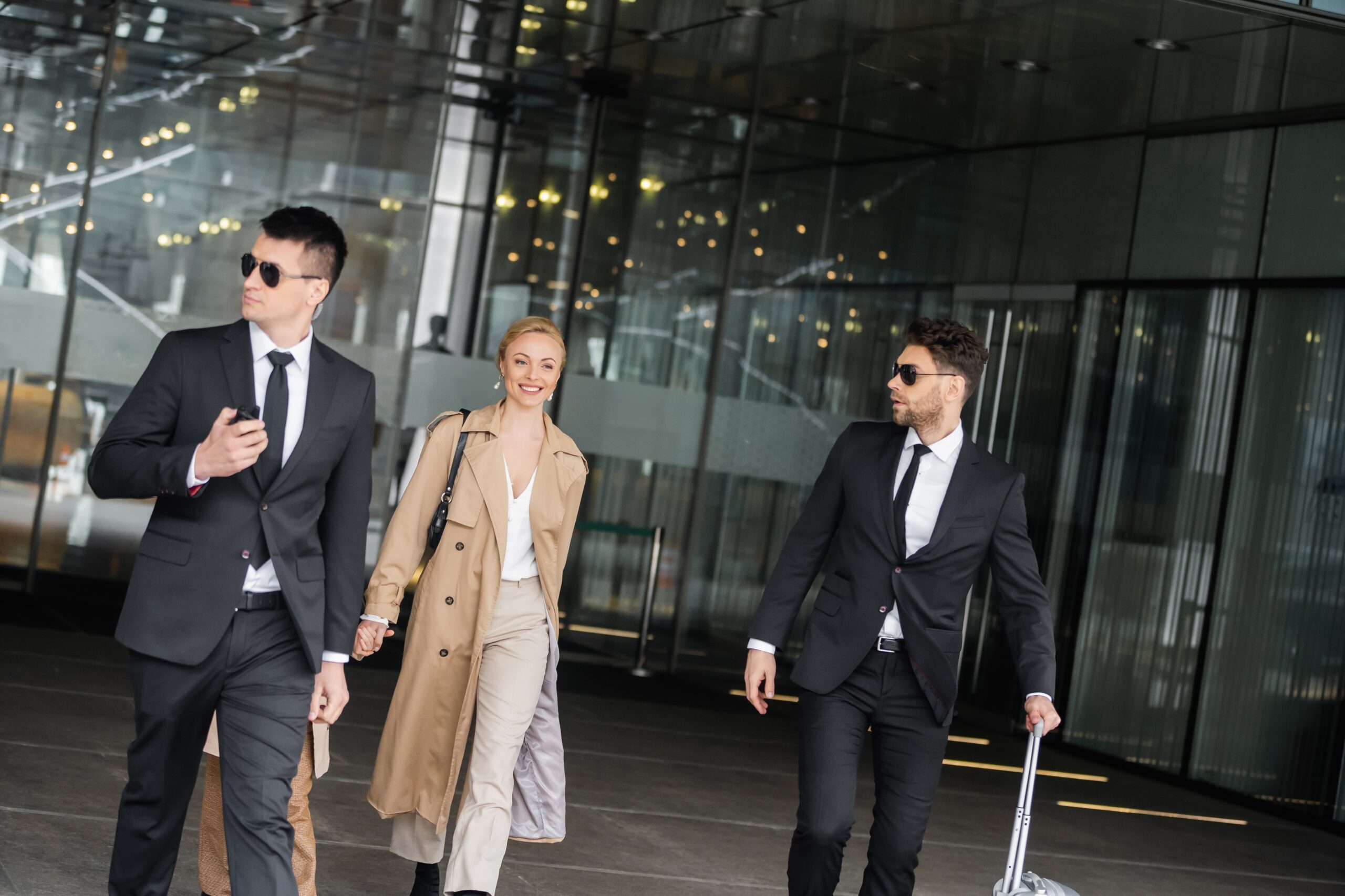 Three people in business attire exit a modern glass building. Two men in black suits and sunglasses walk beside a woman in a beige trench coat. One man is pulling a suitcase. All appear confident and professional.