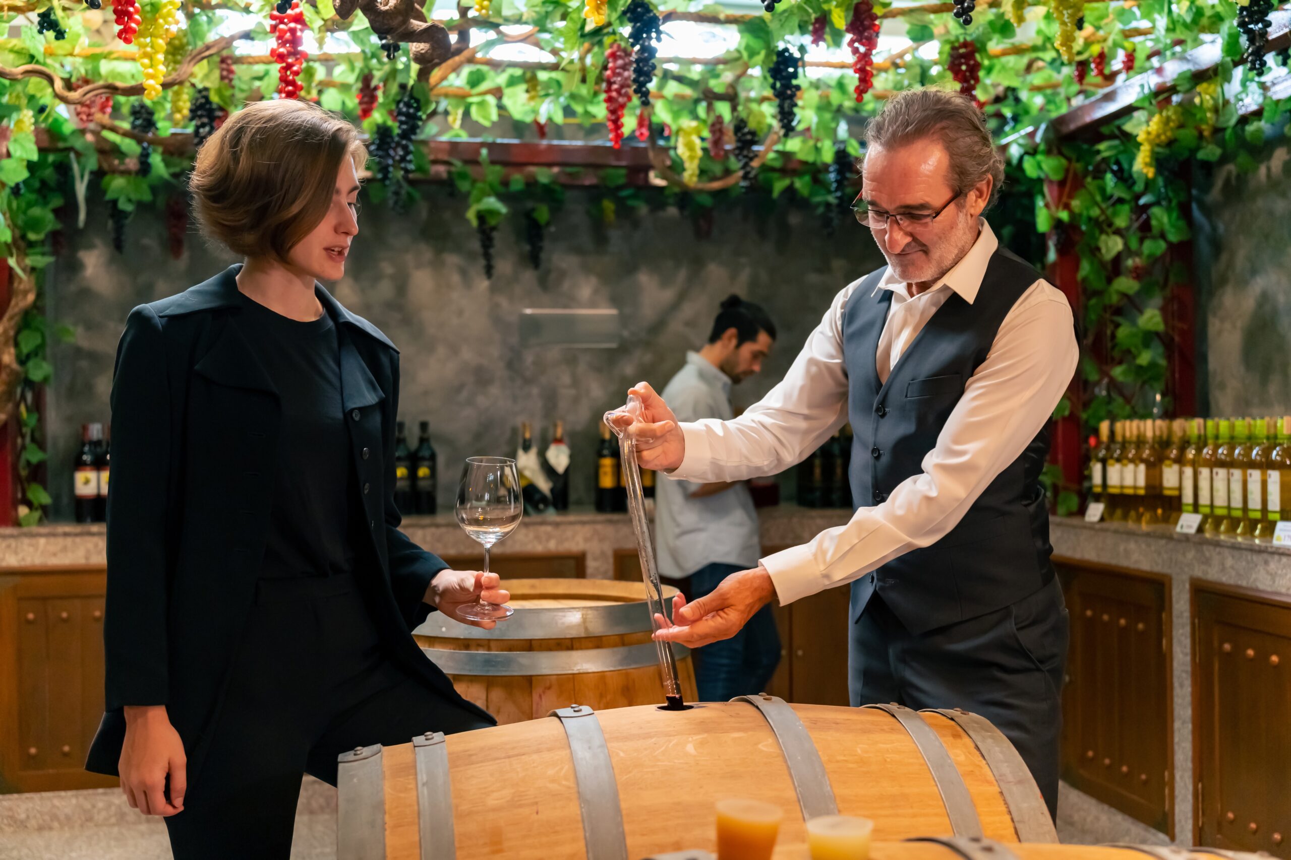 A man pours wine from a barrel into a glass while a woman watches in a winery setting decorated with grapevines and bottles; another man is in the background.